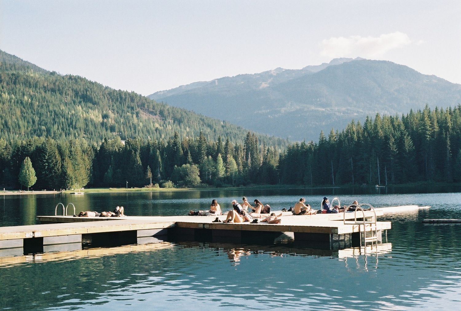 People relaxing on wooden dock platforms at mountain lake surrounded by evergreen forest.