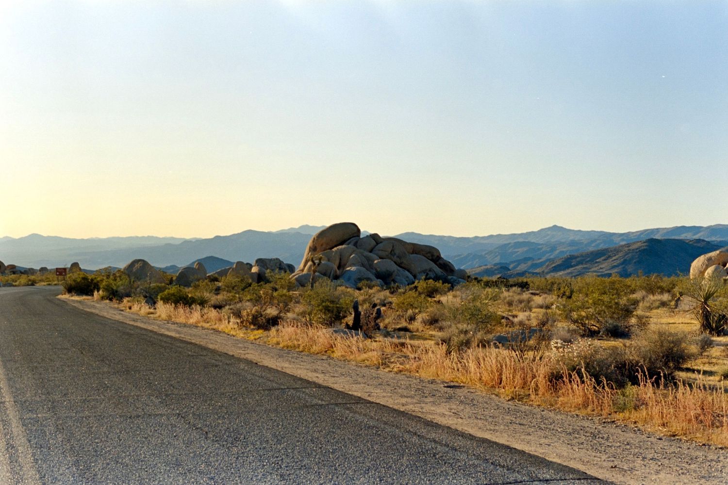 Empty desert road stretches toward distant mountains with large boulder formations.