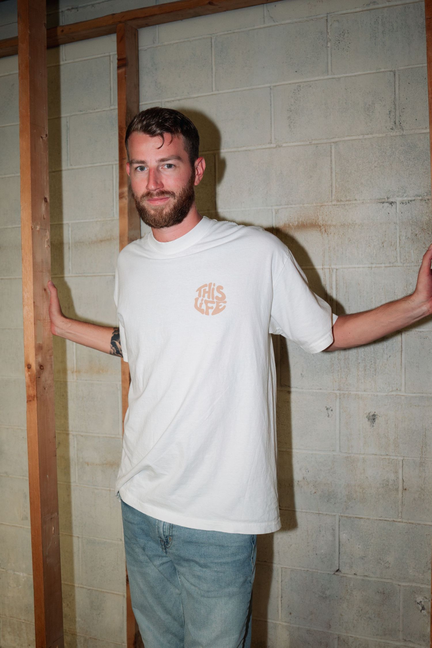 Person in white logo t-shirt poses against concrete wall in basement setting.
