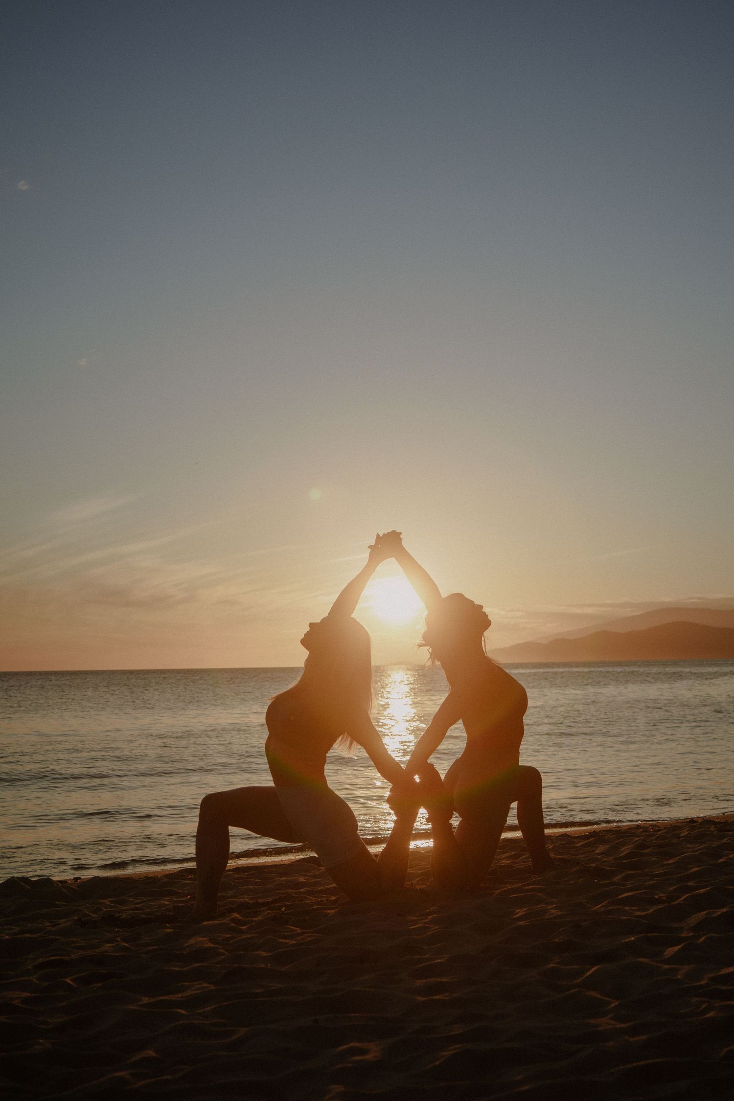 Two silhouettes perform synchronized yoga poses on beach at golden hour.