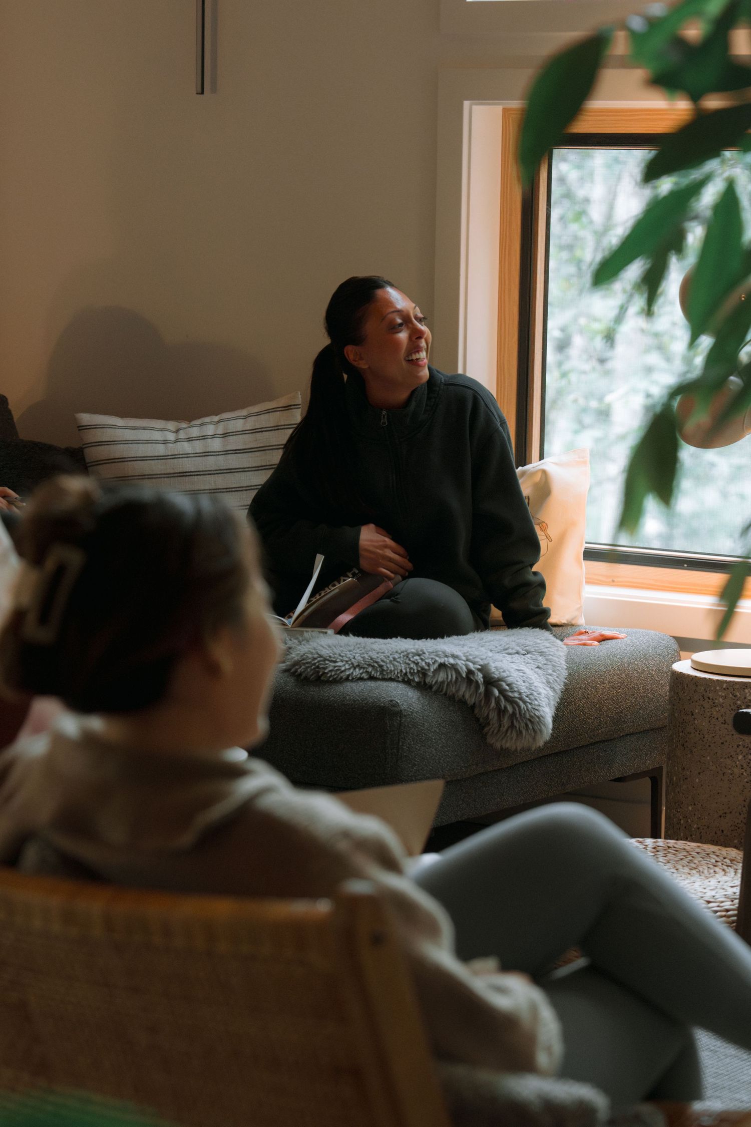 People relaxing in a sunlit room with large windows and indoor plants.
