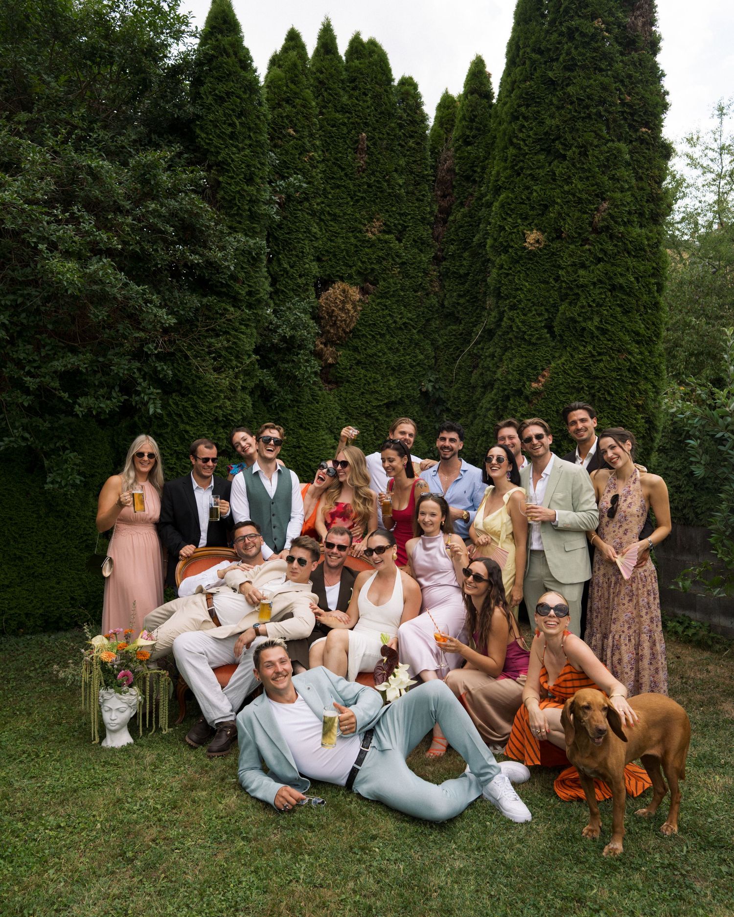 Large group of stylish friends posing together outdoors on green lawn with tall evergreen trees backdrop.