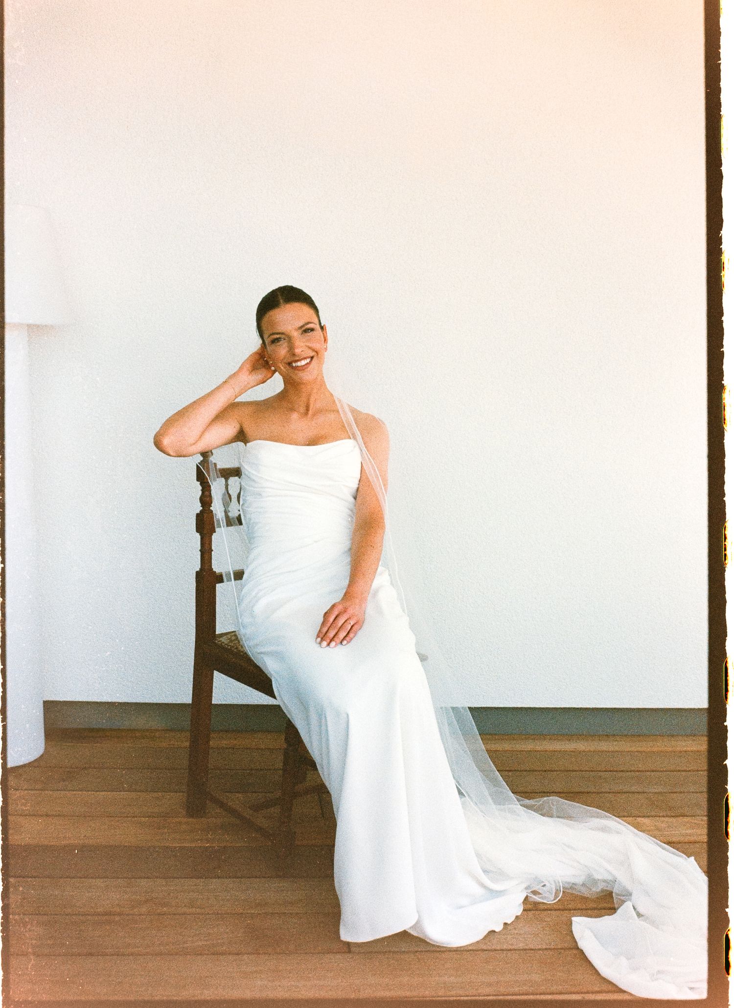 Smiling bride in a white halter wedding gown with veil train, seated on a wooden chair against a neutral wall.