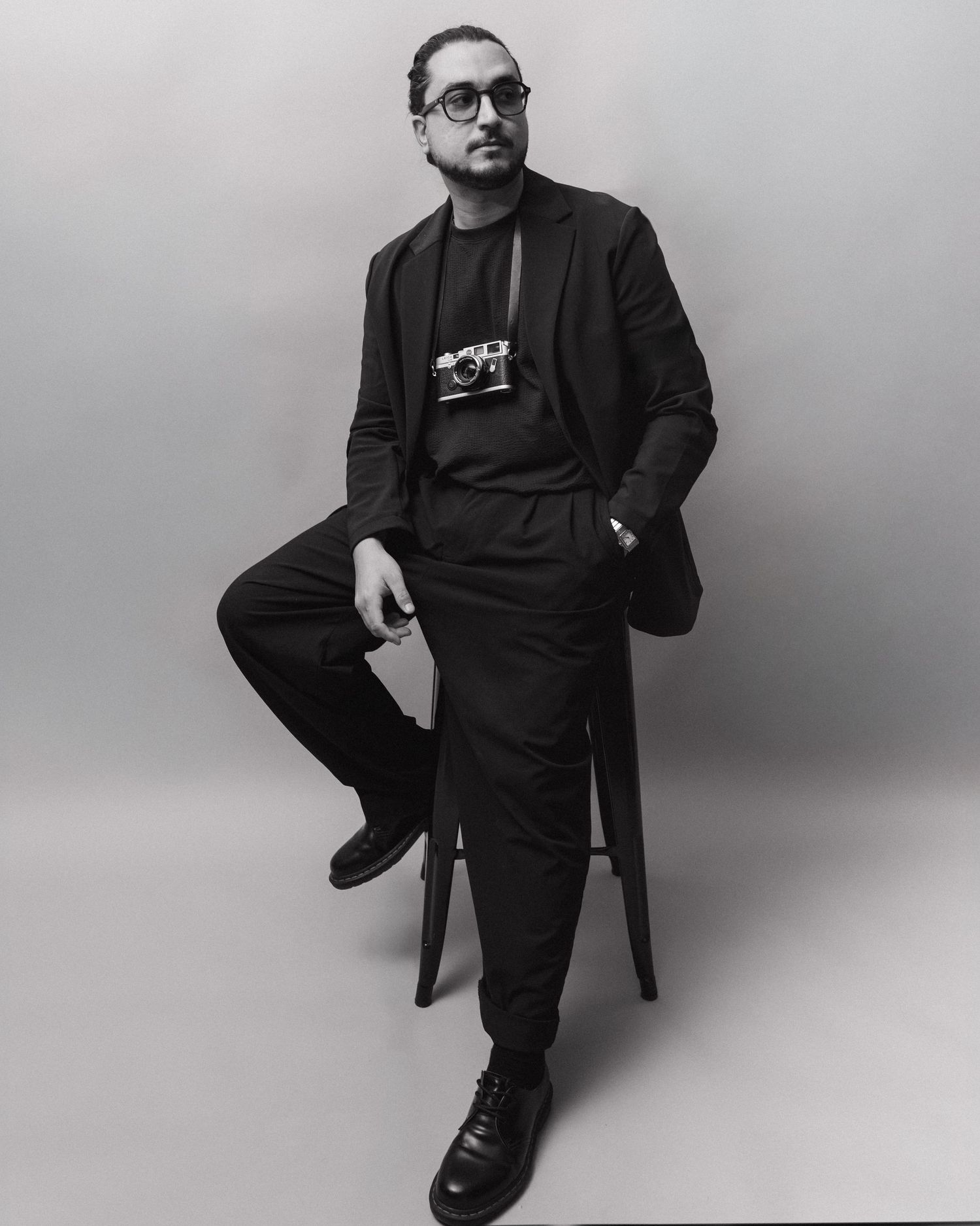 Black and white portrait of someone in a dark suit and glasses sitting on a stool against a plain backdrop.