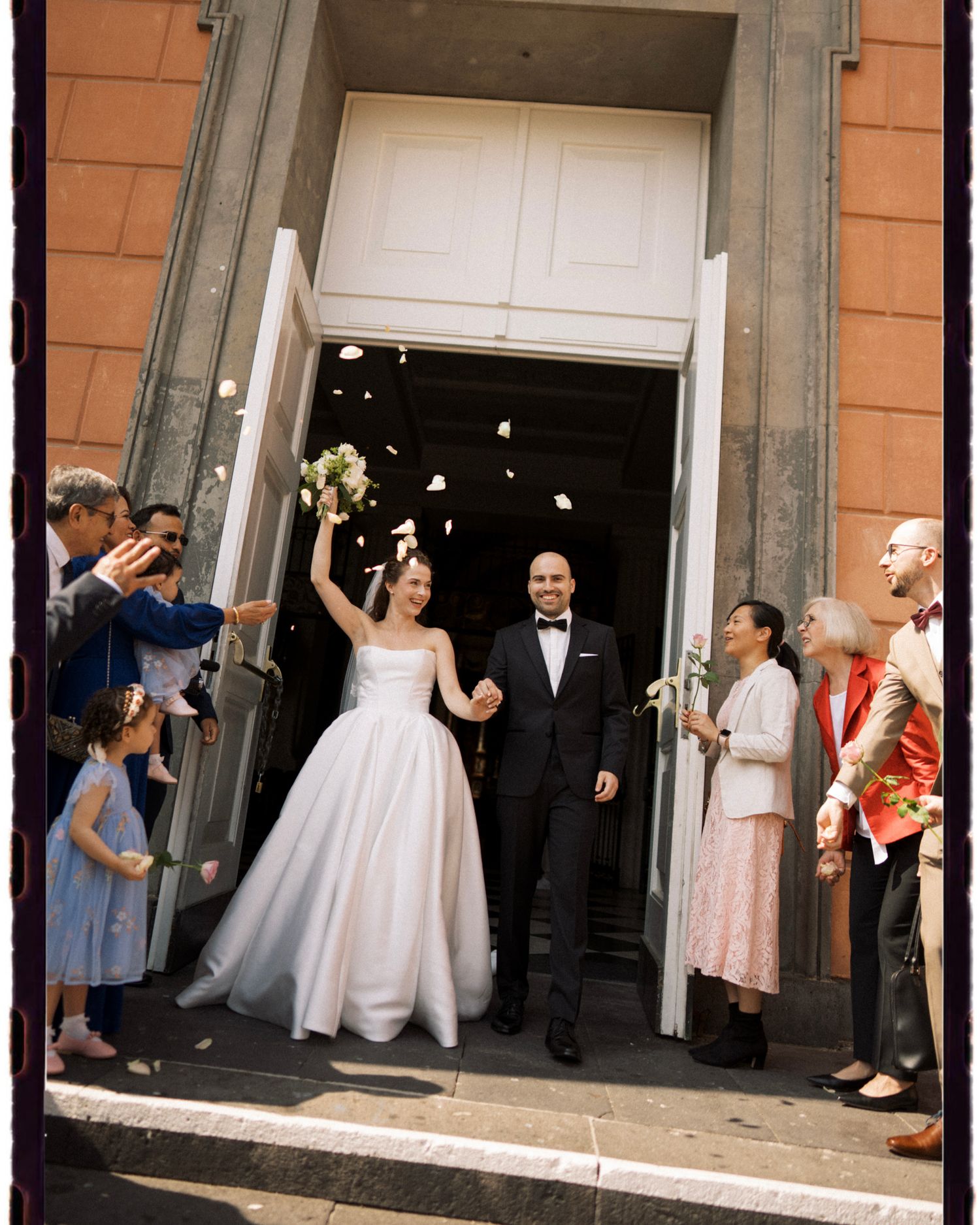 Bride and groom exit church doors as guests toss flower petals in joyful wedding celebration.