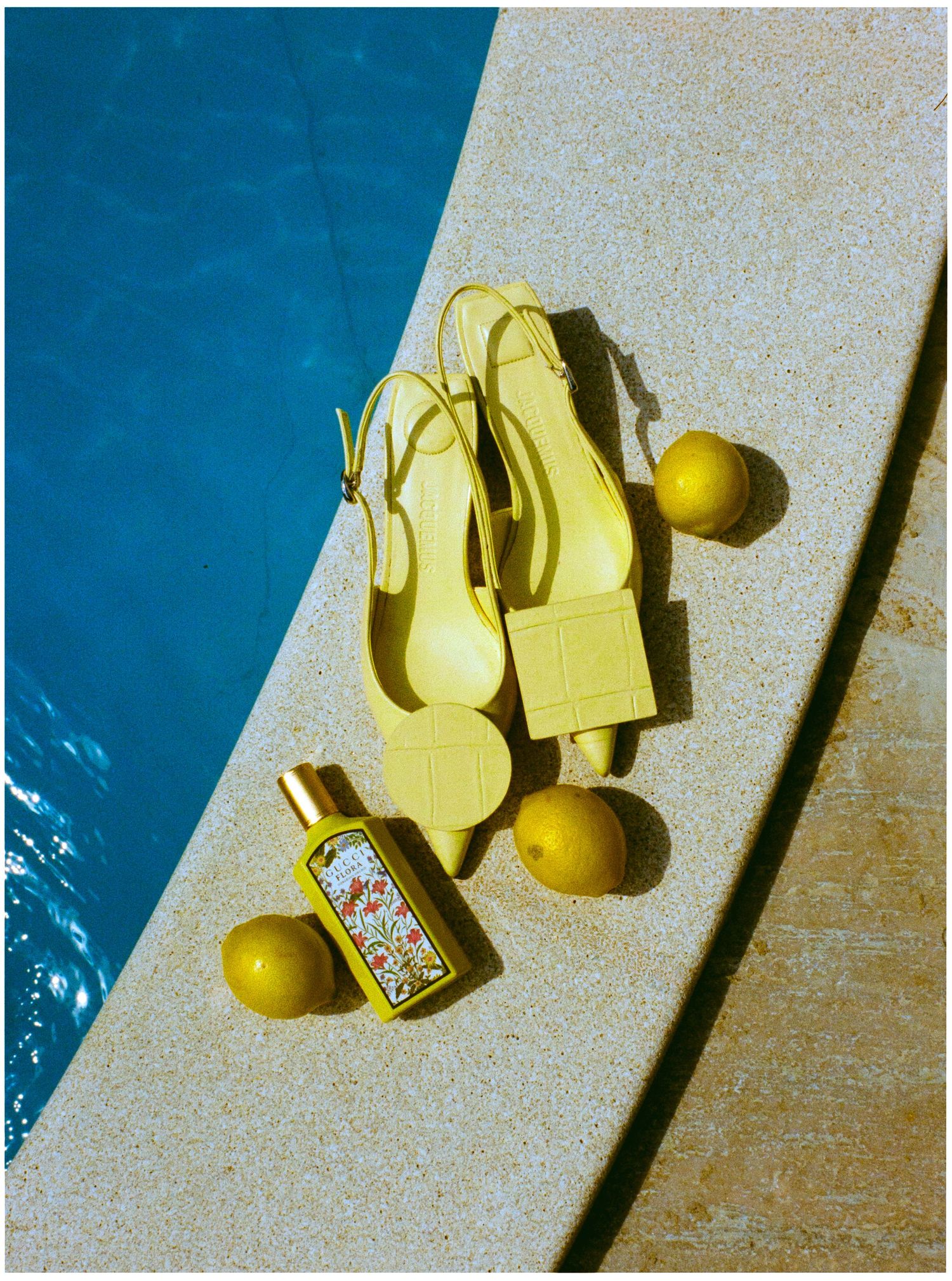 Yellow slingback heels and floral perfume bottle beside lemons on a sun-drenched poolside ledge.
