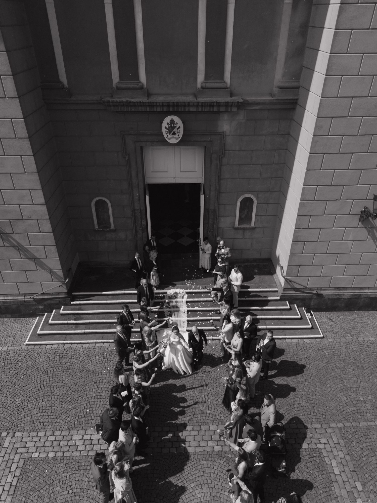 Black and white aerial view of a wedding ceremony outside a classic church with guests lining the steps.