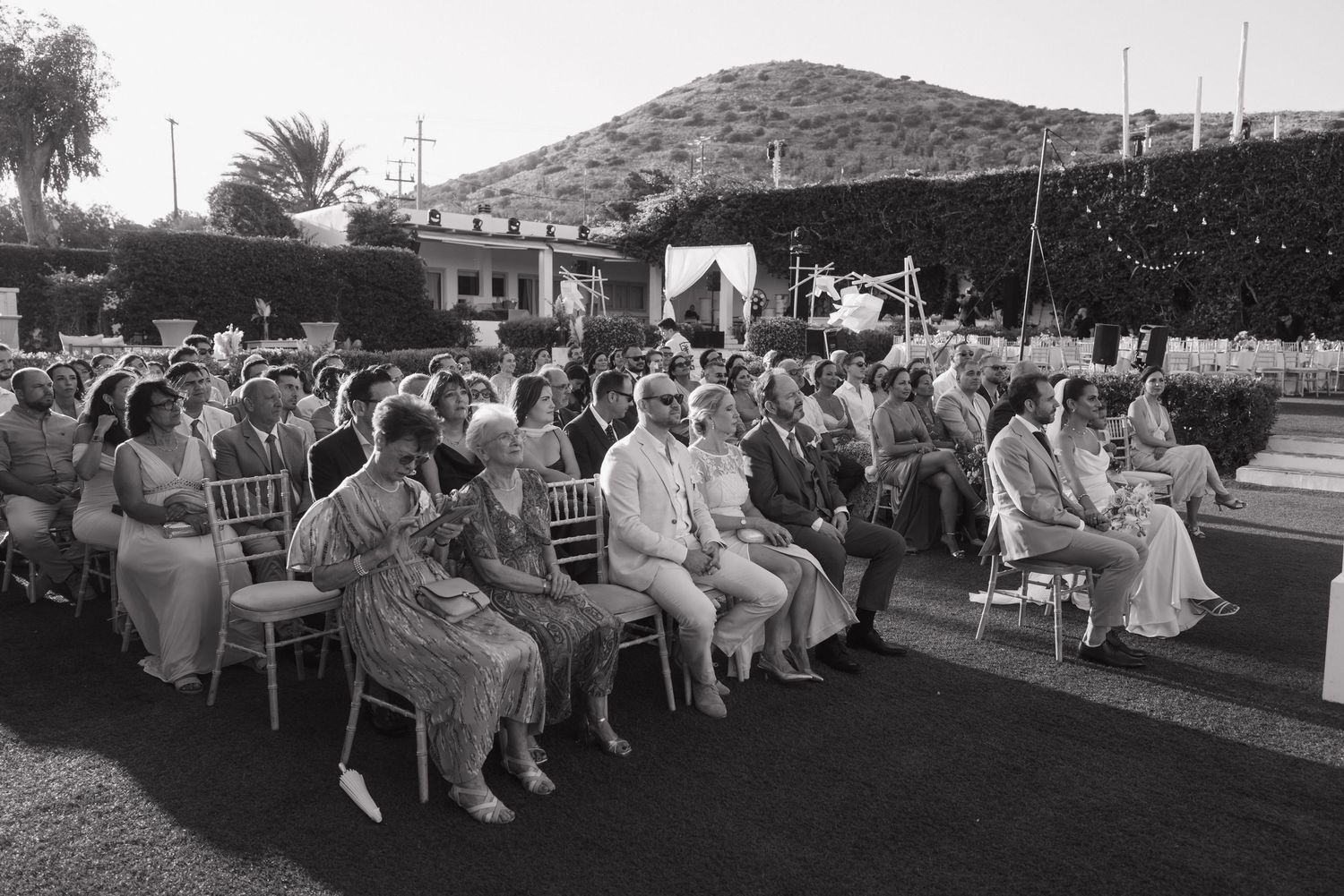 A panoramic view shows a large crowd seated outdoors at an evening event with palm trees in the background.