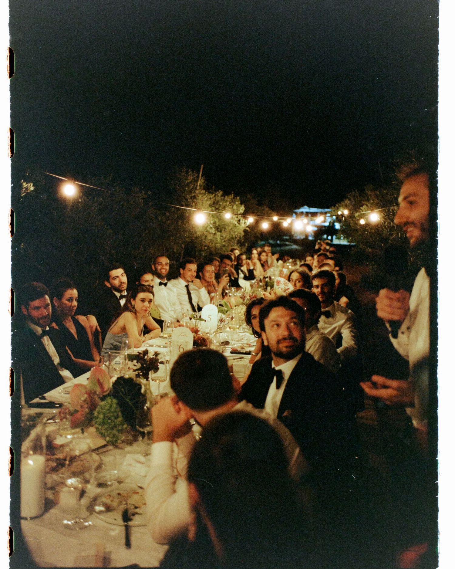 Elegant outdoor wedding reception dinner at night with string lights, guests in formal attire seated at a long decorated table.
