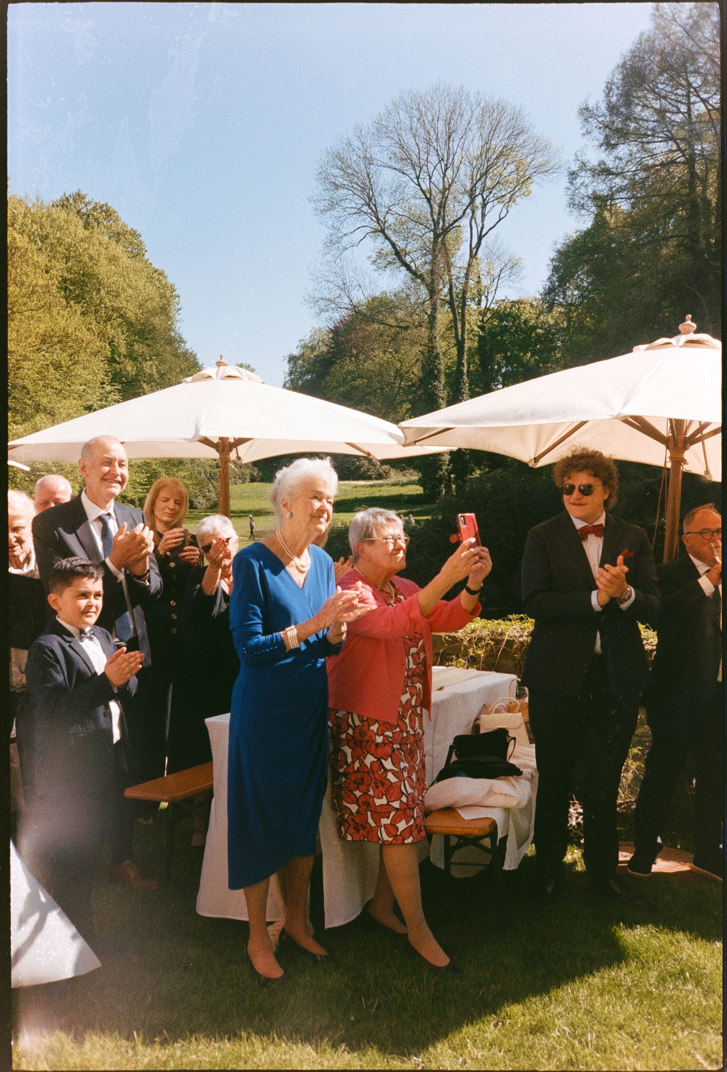 Elderly women dancing outdoors at a garden party while guests clap and take photos under white umbrellas.