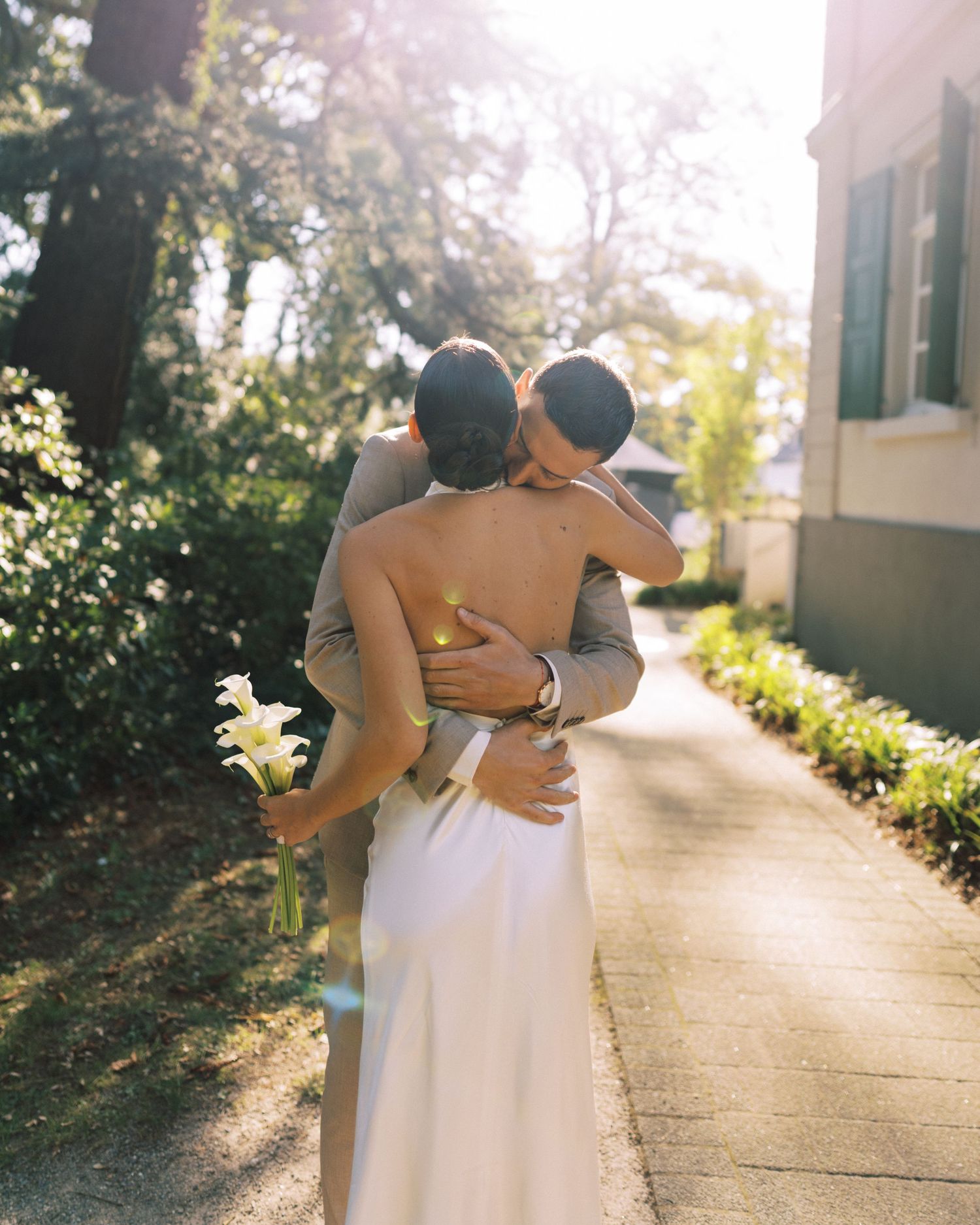 A couple embraces in warm sunlight on a garden pathway creating a romantic silhouette against the bright background.