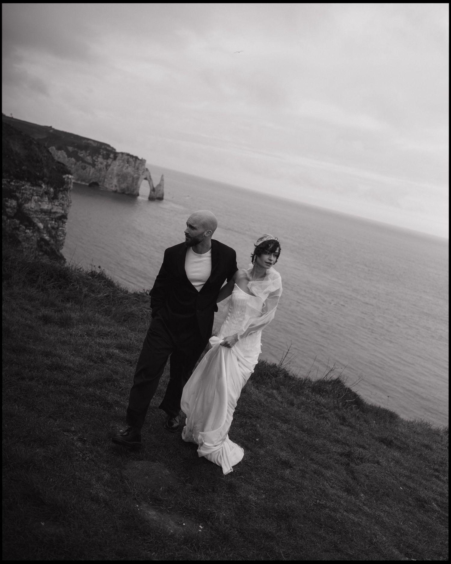 Black and white photo of a couple in wedding attire posing on a cliff overlooking the ocean with dramatic rock formations.