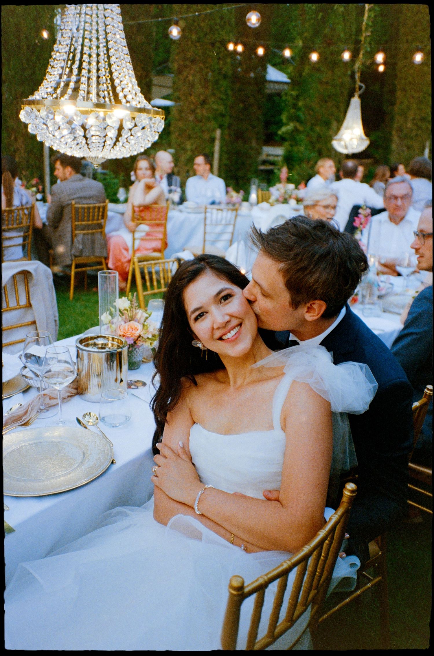 Crystal chandeliers hang above elegant outdoor wedding dinner tables set with white linens and place settings at dusk.