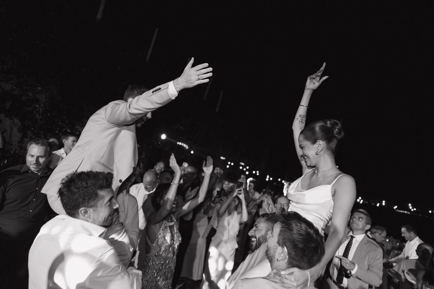Black and white wedding reception photo of bride and groom dancing joyfully, lifted by guests at an outdoor celebration.