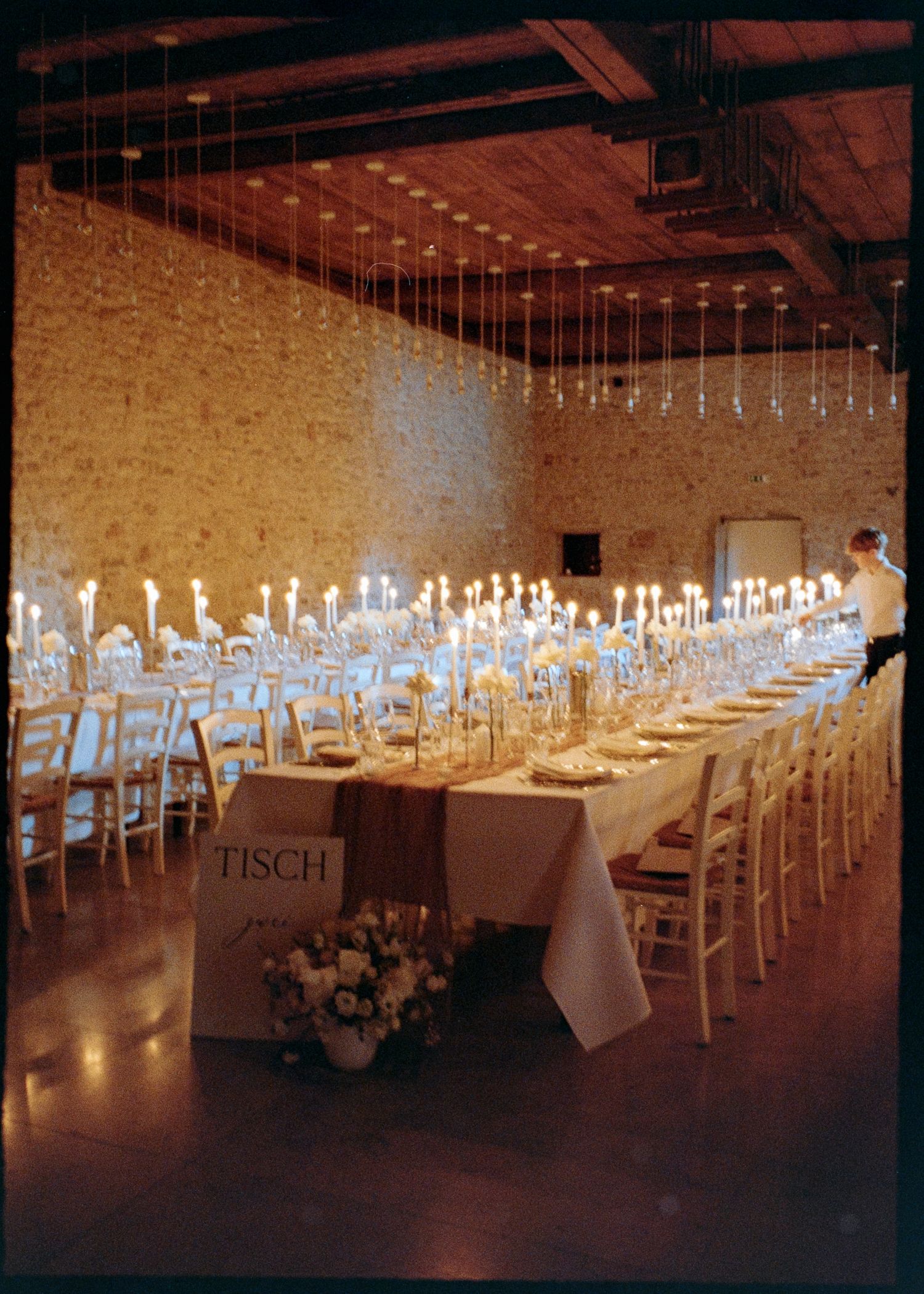 Long white banquet tables with elegant place settings and hanging lights create a dramatic reception space in a dark venue.
