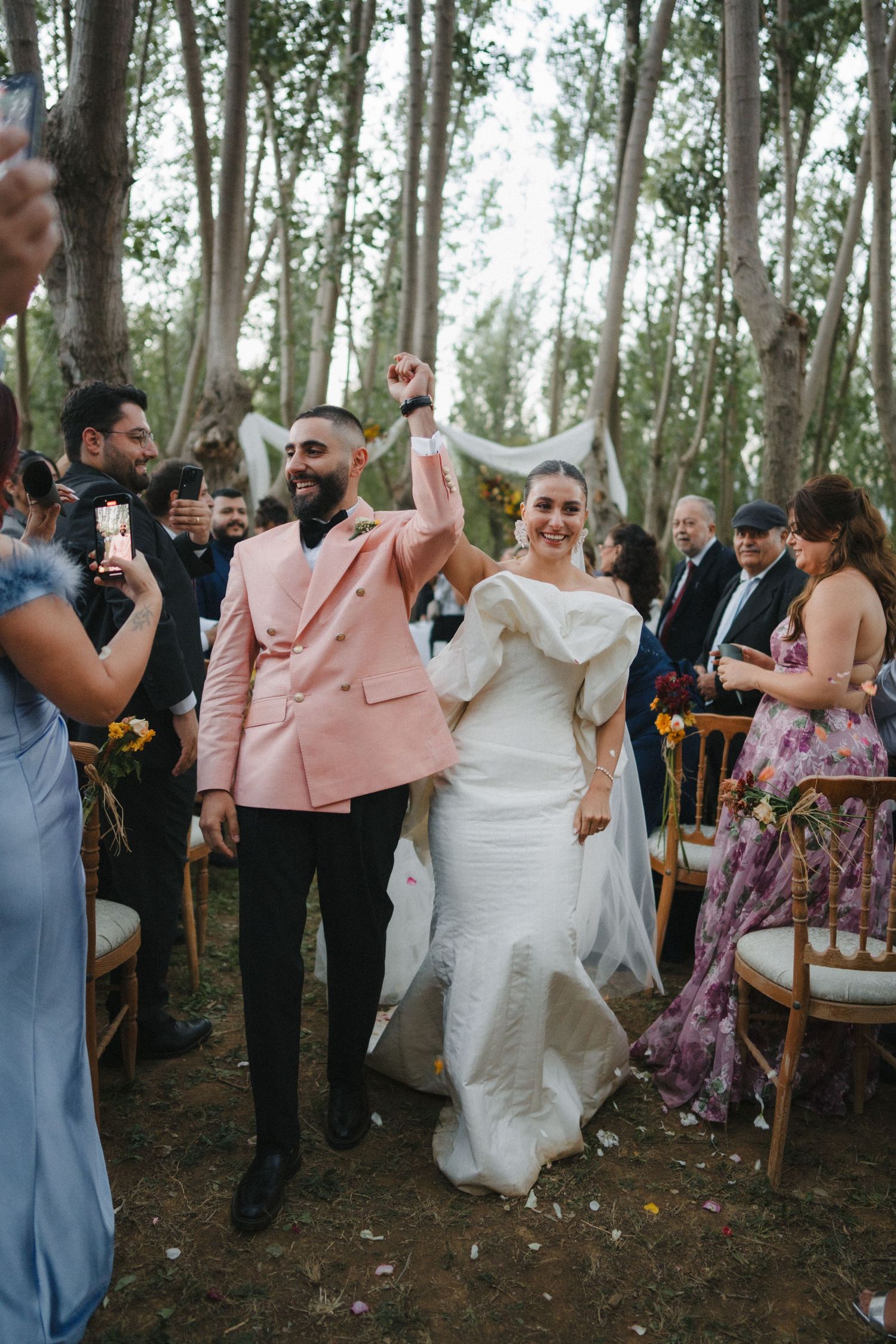 Wedding guests celebrate joyfully in a wooded outdoor ceremony setting with chairs arranged among trees.