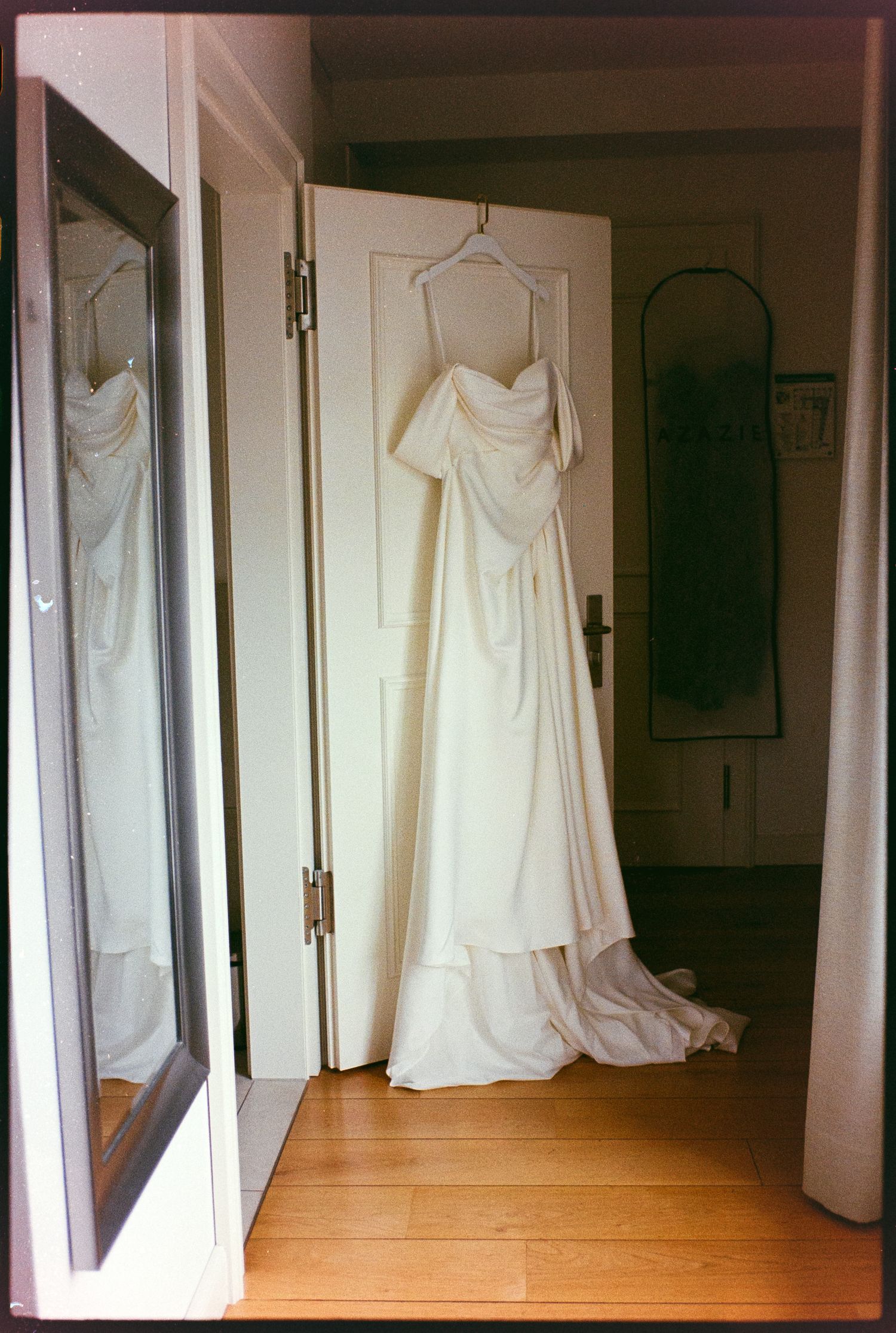 Elegant white wedding gown with off-shoulder design hanging on a door in a warmly lit room with hardwood floors.
