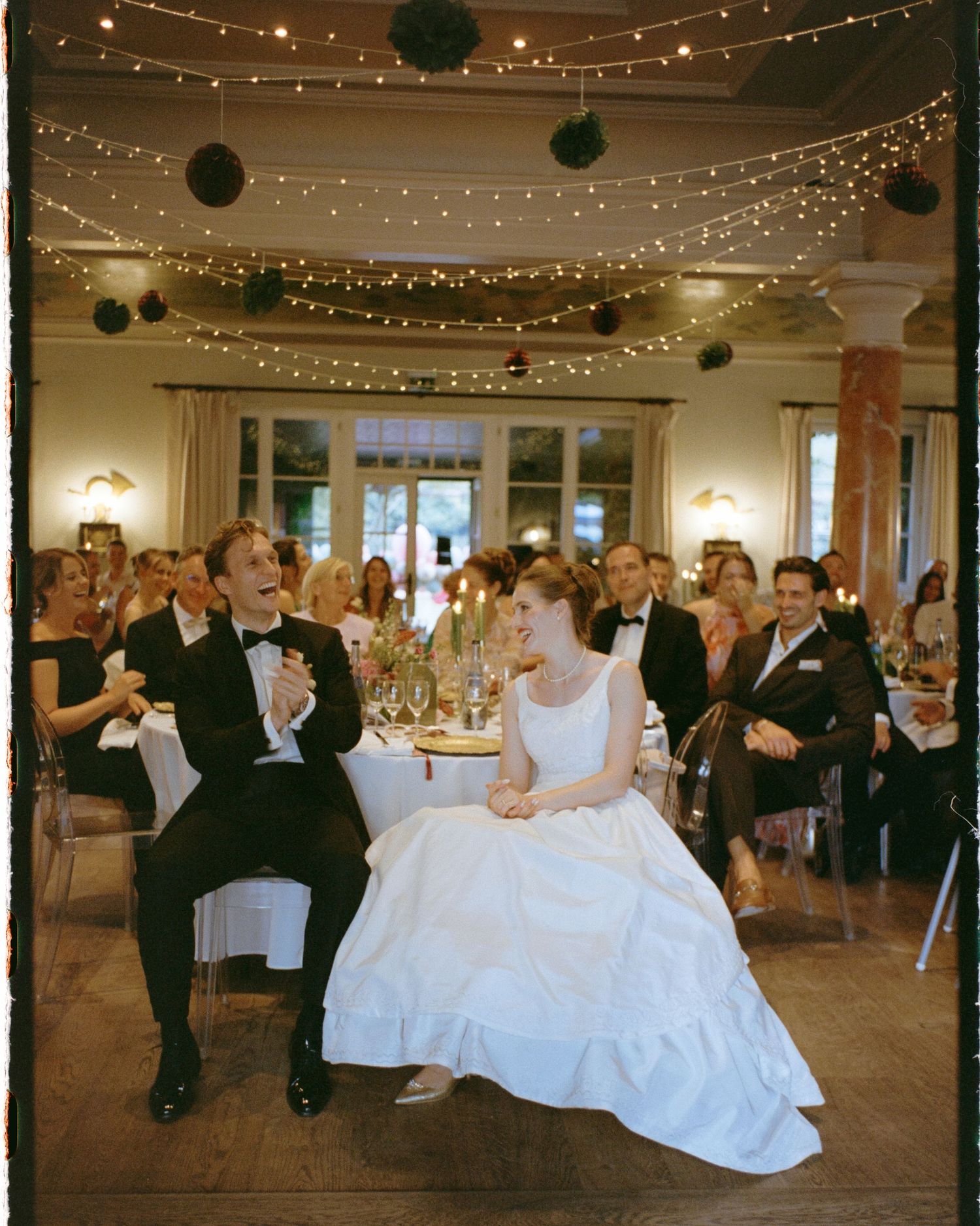 Bride and groom sit together at wedding reception table, surrounded by guests under warm string lights and floral decor.