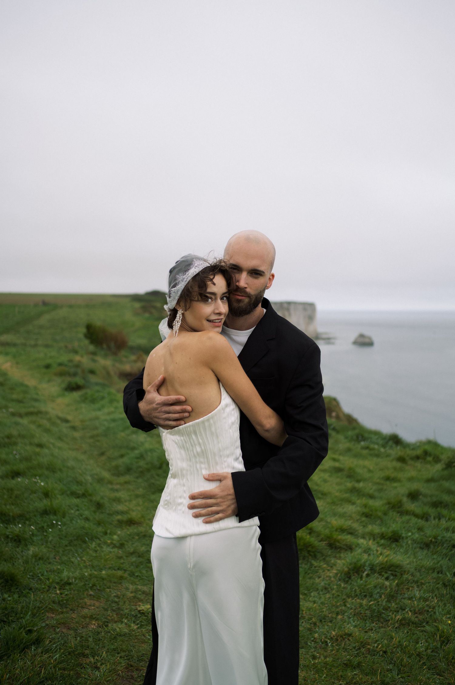 Bride and groom embrace on coastal cliffside, she in strapless white gown with veil, he in black suit, ocean backdrop.