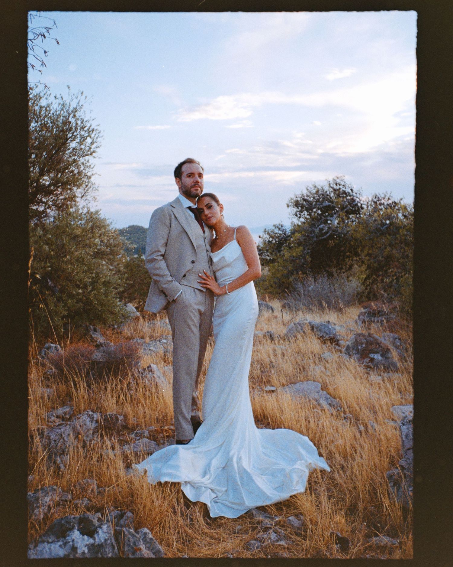 A couple in wedding attire embraces in a golden field of tall grass during sunset.