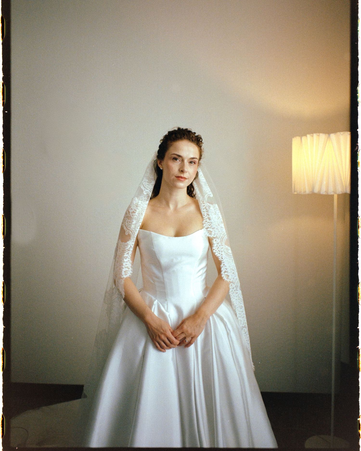 A bride in a white satin ballgown and lace veil stands elegantly before a softly lit floor lamp.