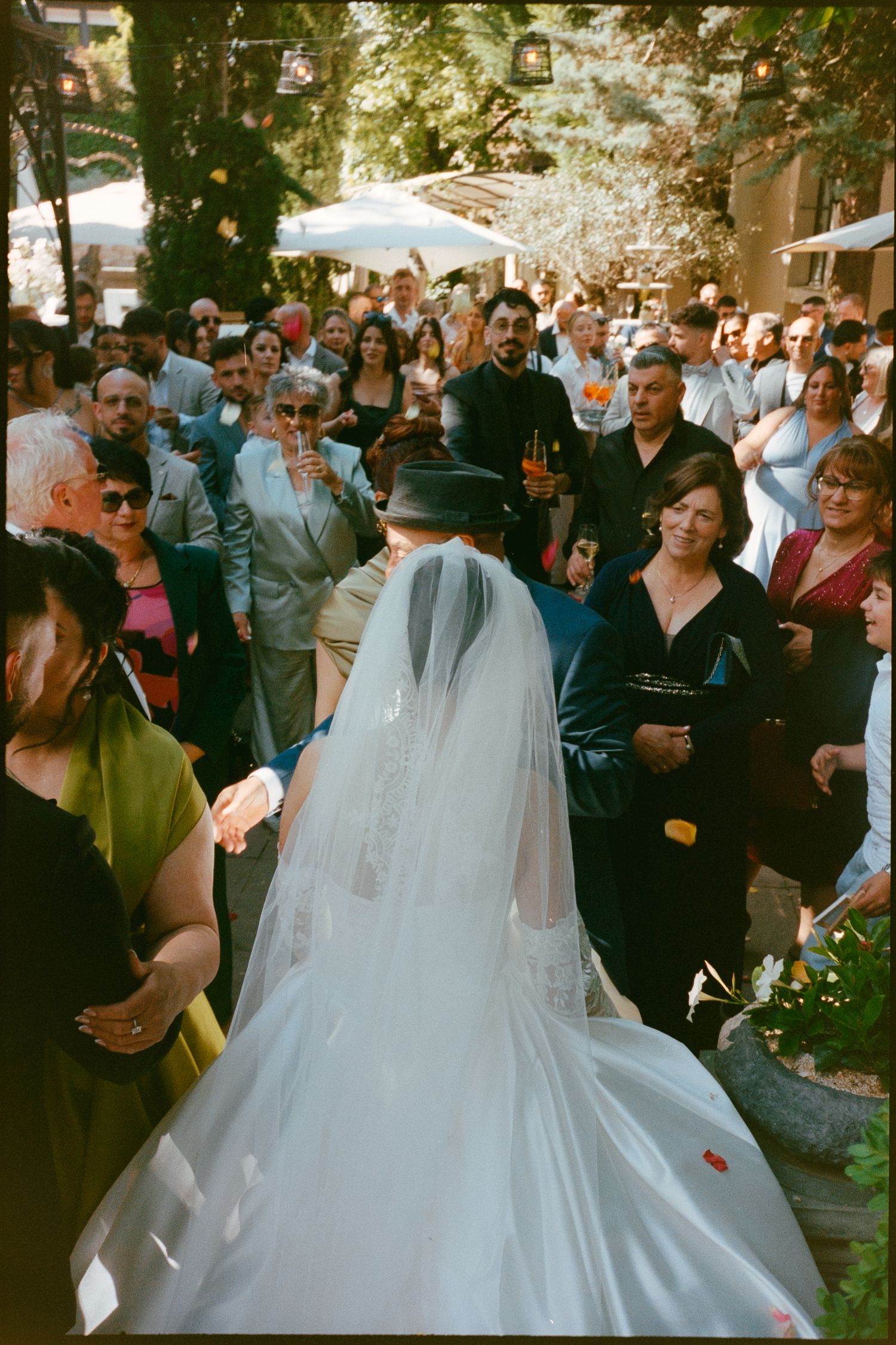 Bride in white gown and veil walks down outdoor aisle surrounded by wedding guests in a garden setting.