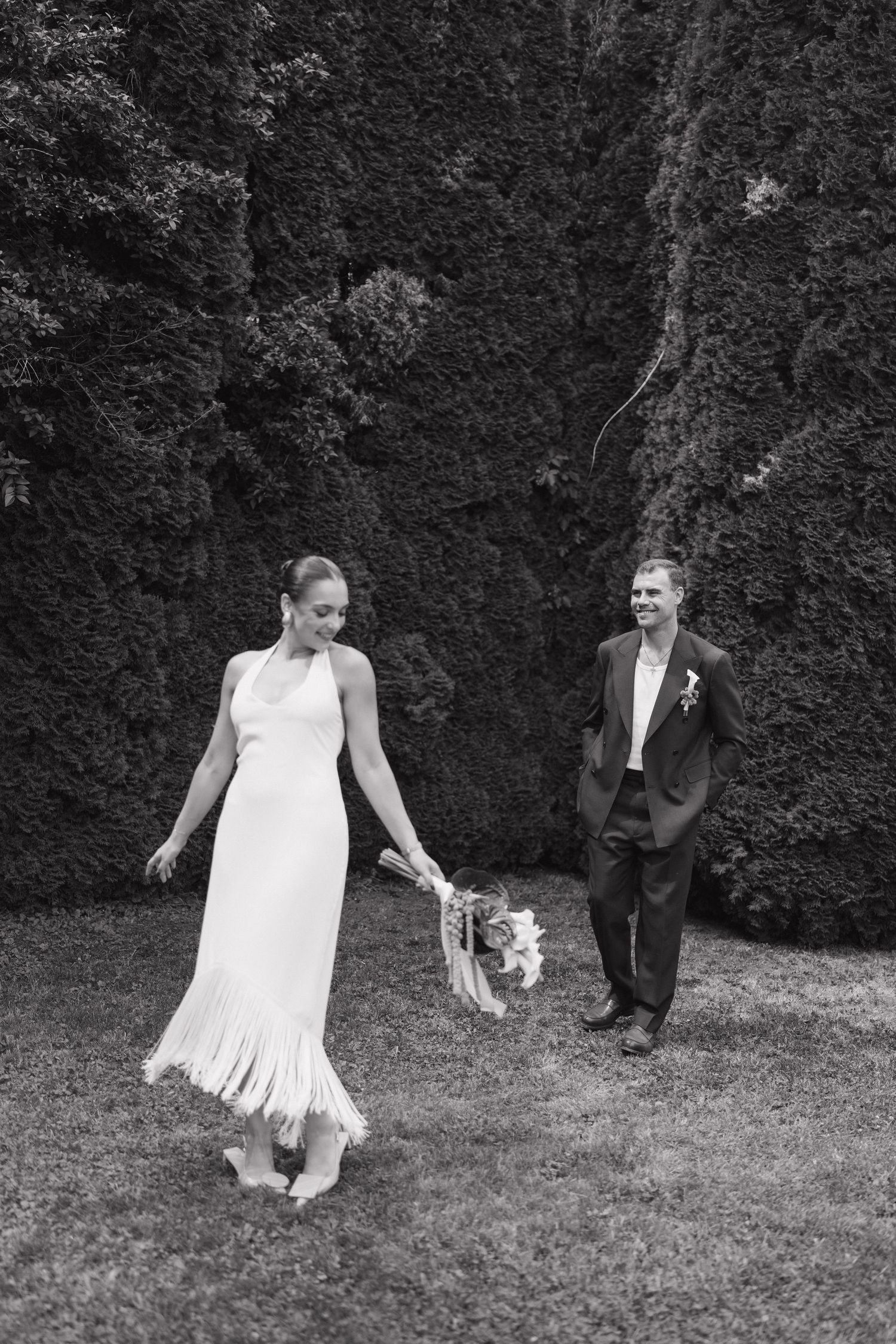 Black and white photo of a bride in white fringed dress and groom in suit walking on grass before ivy wall.