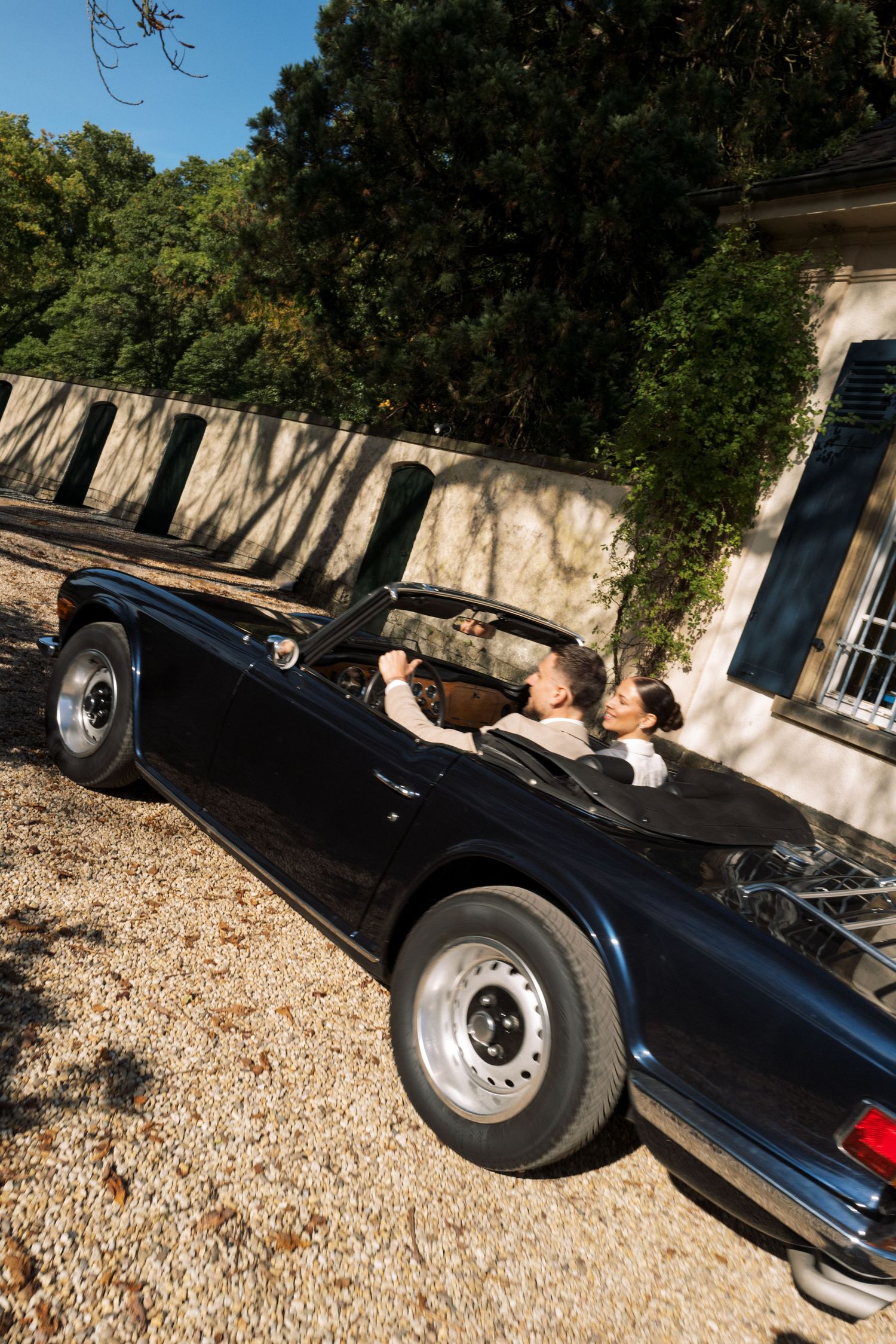 A black classic convertible car parked on gravel with trees and a building in the background.