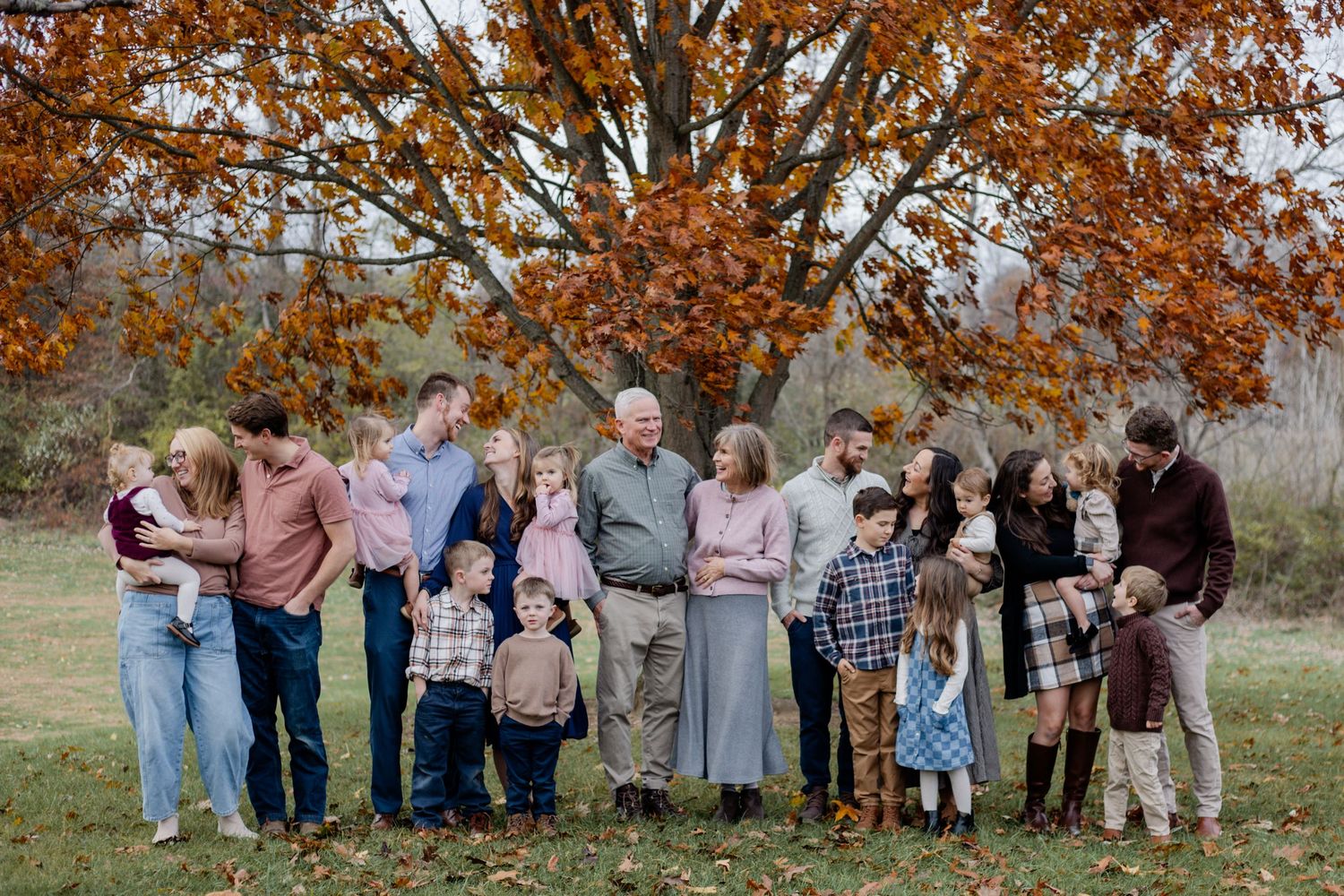 Large multigenerational family portrait outdoors in autumn, gathered under a vibrant orange tree on a green lawn.