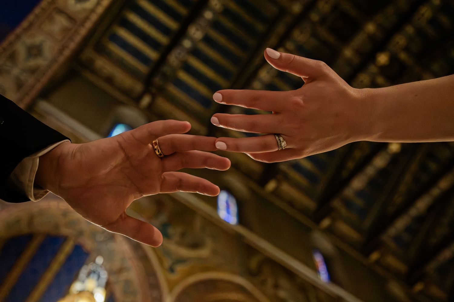 Two hands reaching toward each other with wedding rings in an ornate church setting.