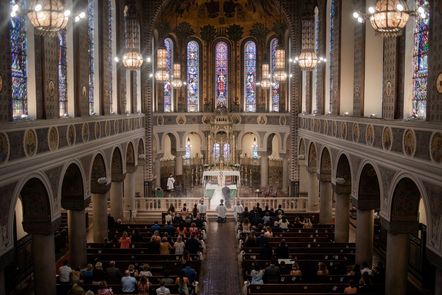 Grand cathedral interior with ornate arches, stained glass windows, and wedding ceremony guests seated in wooden pews.