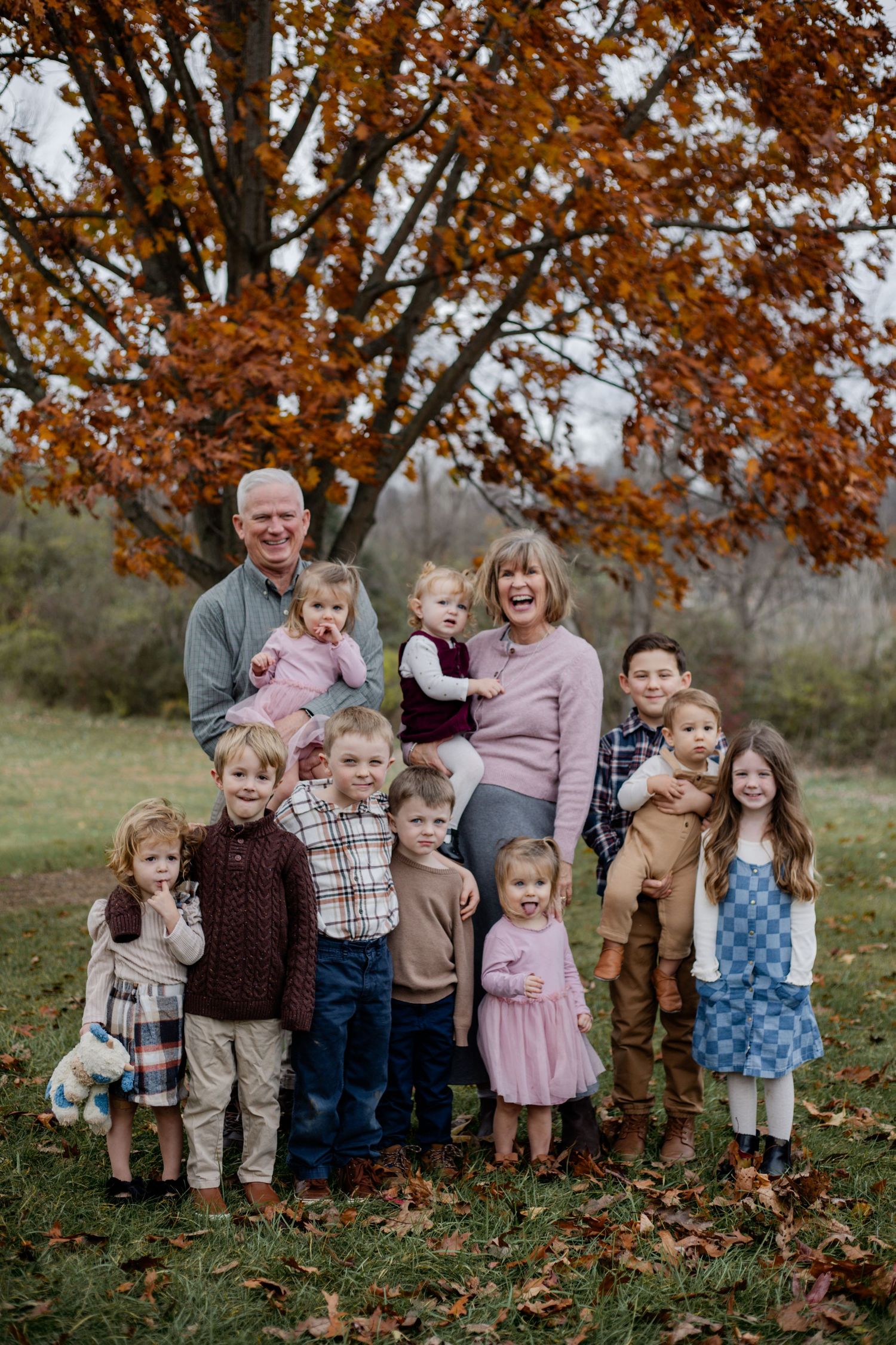Multi-generational family portrait outdoors in autumn, grandparents surrounded by grandchildren under colorful fall foliage.