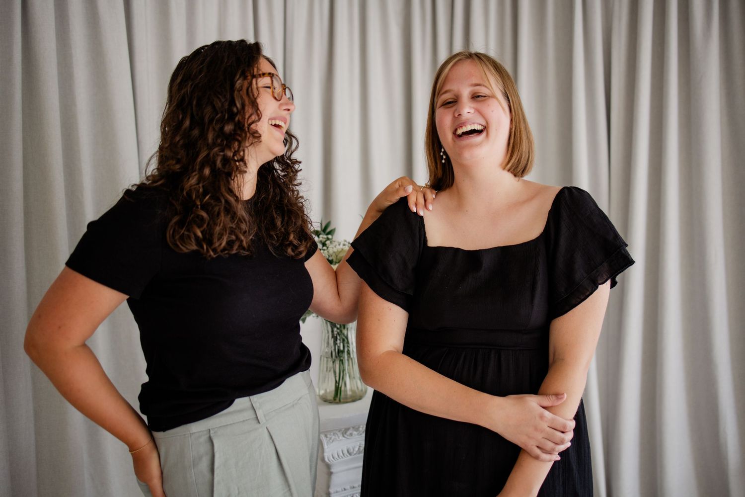 Two women in black tops laugh together against a white curtain backdrop in a bright, elegant setting.