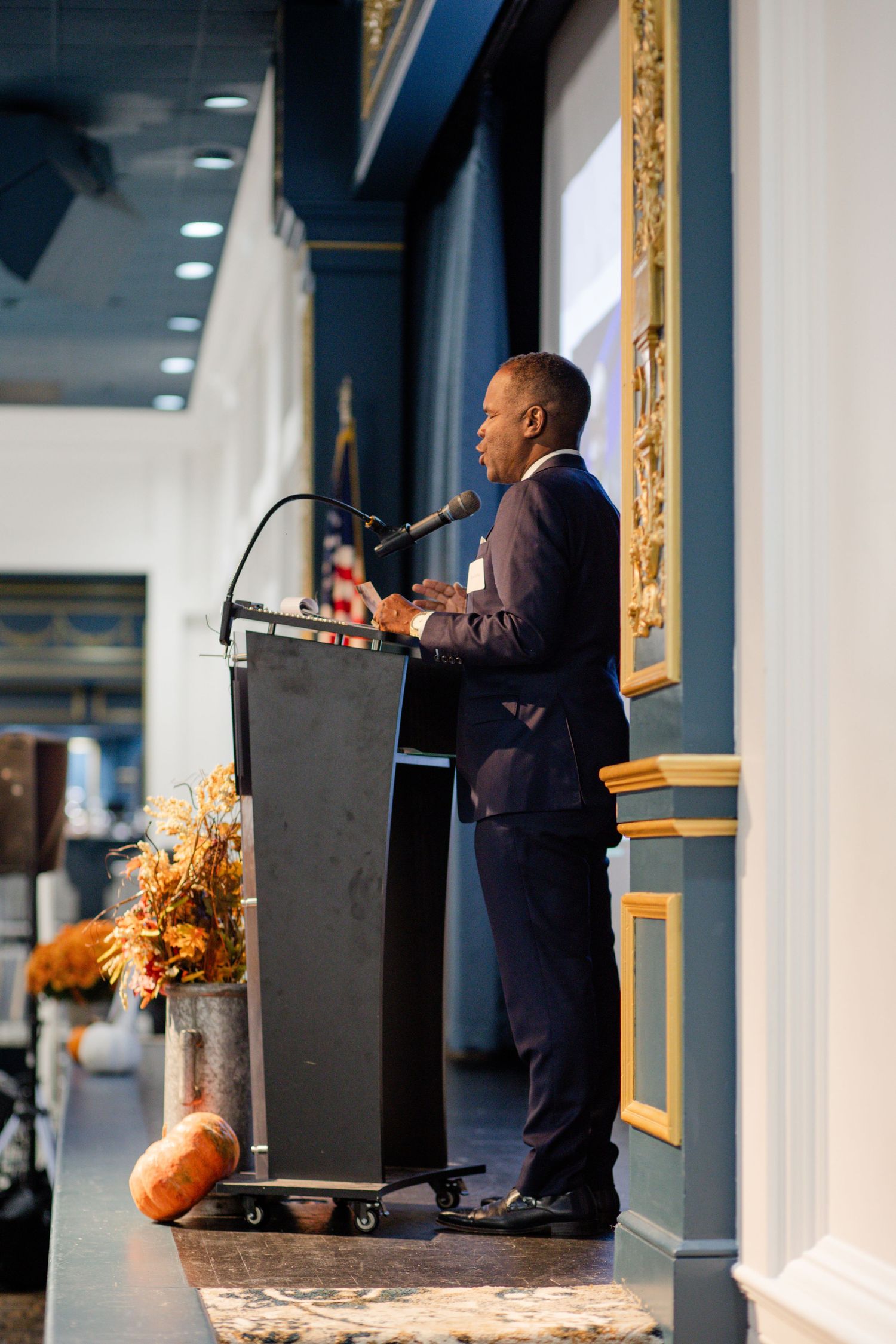 Man in navy suit speaking at a black podium with microphone, flanked by autumn floral arrangements in an elegant venue.
