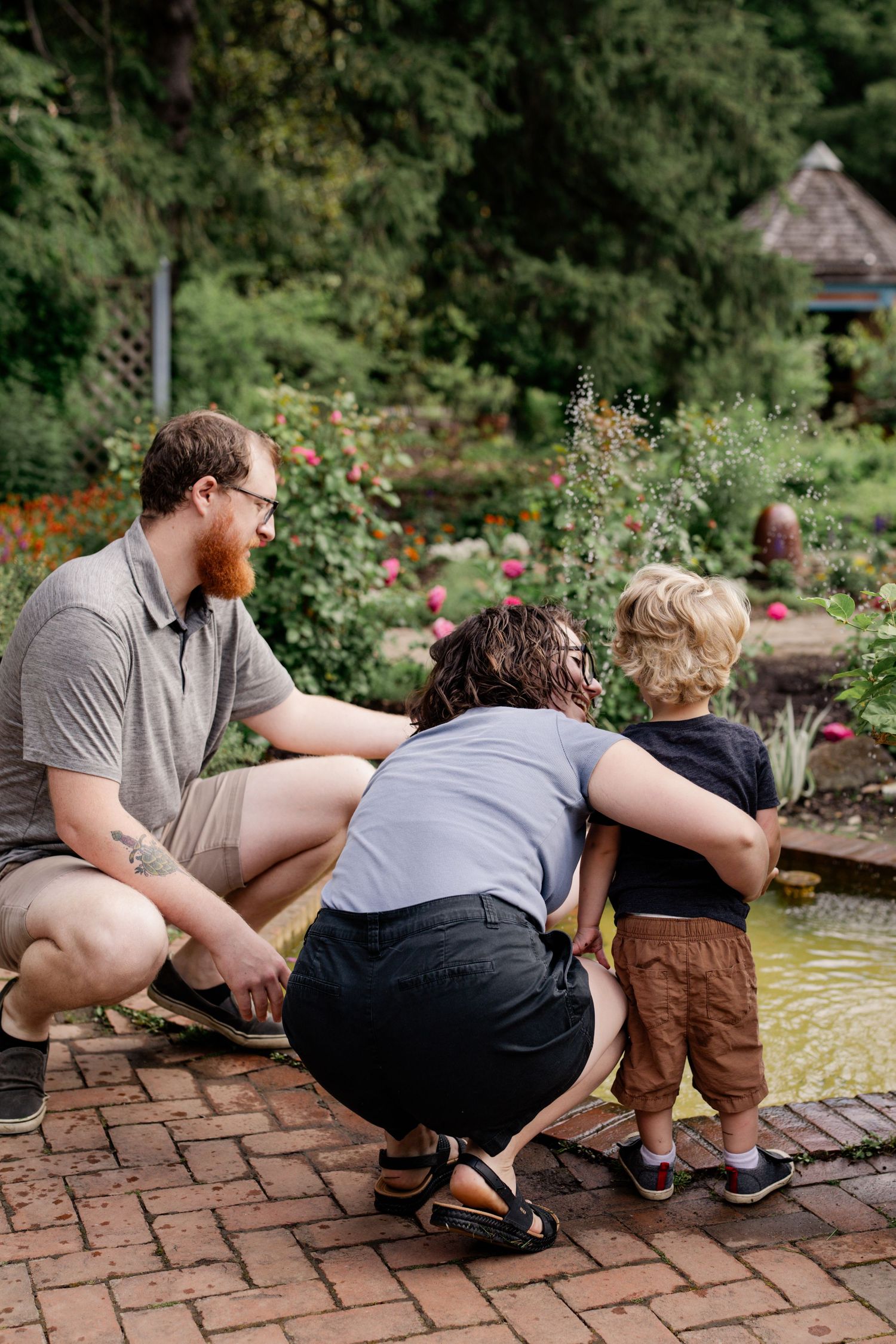 Family with toddler crouching by a garden pond surrounded by lush greenery and blooming flowers.