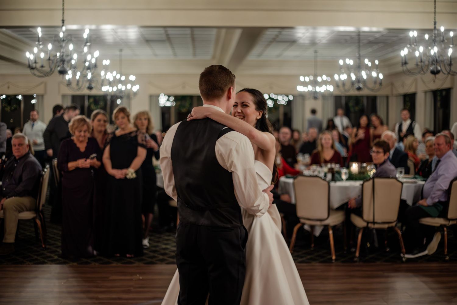 Bride and groom share their first dance at elegant wedding reception with guests watching in candlelit ballroom.