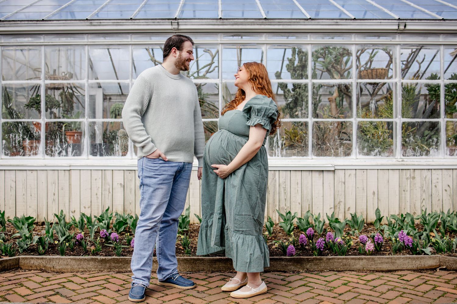 Expecting couple stands facing each other outdoors near a greenhouse, woman in green maternity dress cradling her baby bump.