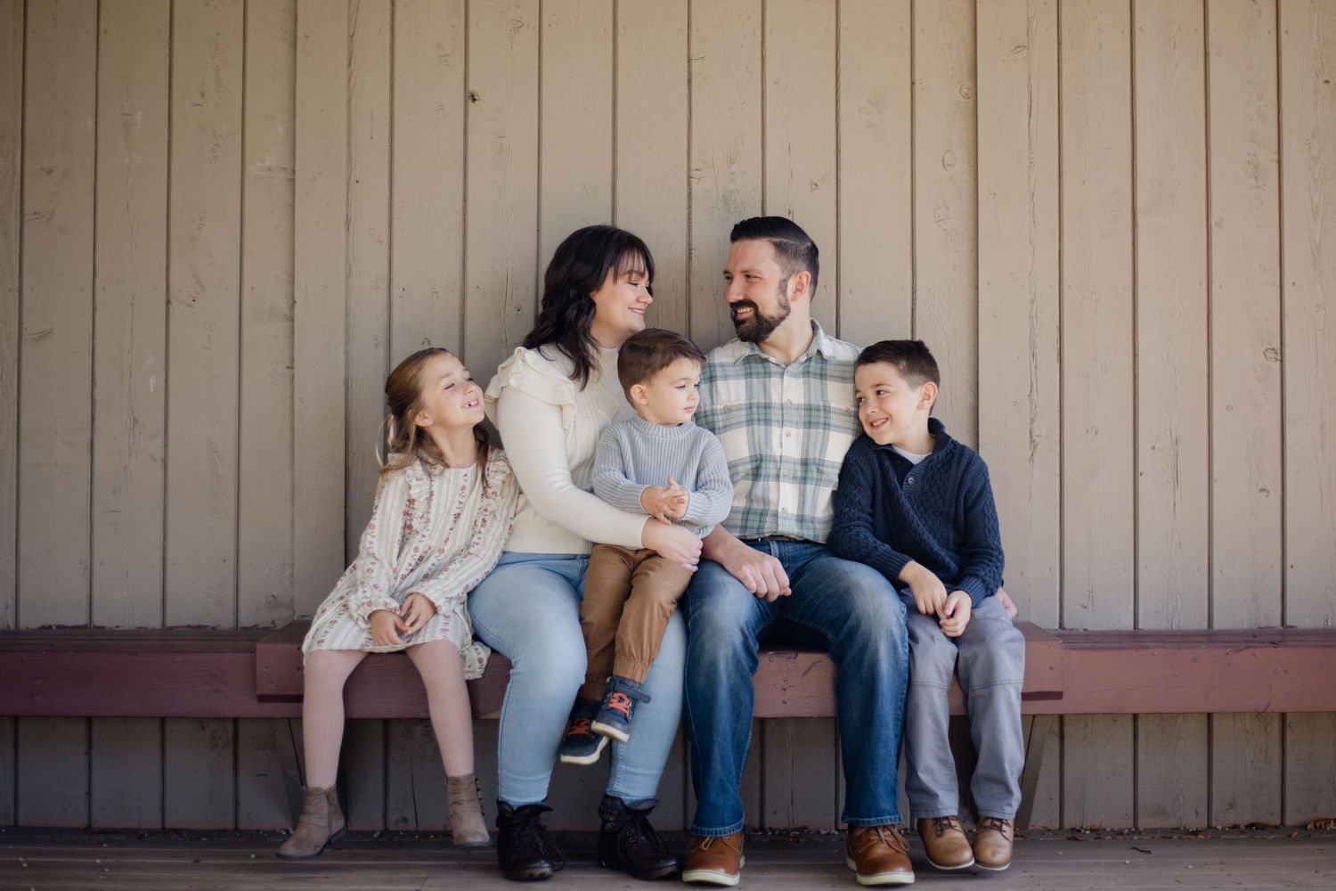 A happy family of five sits together on a bench, smiling and interacting outdoors near a wooden wall.