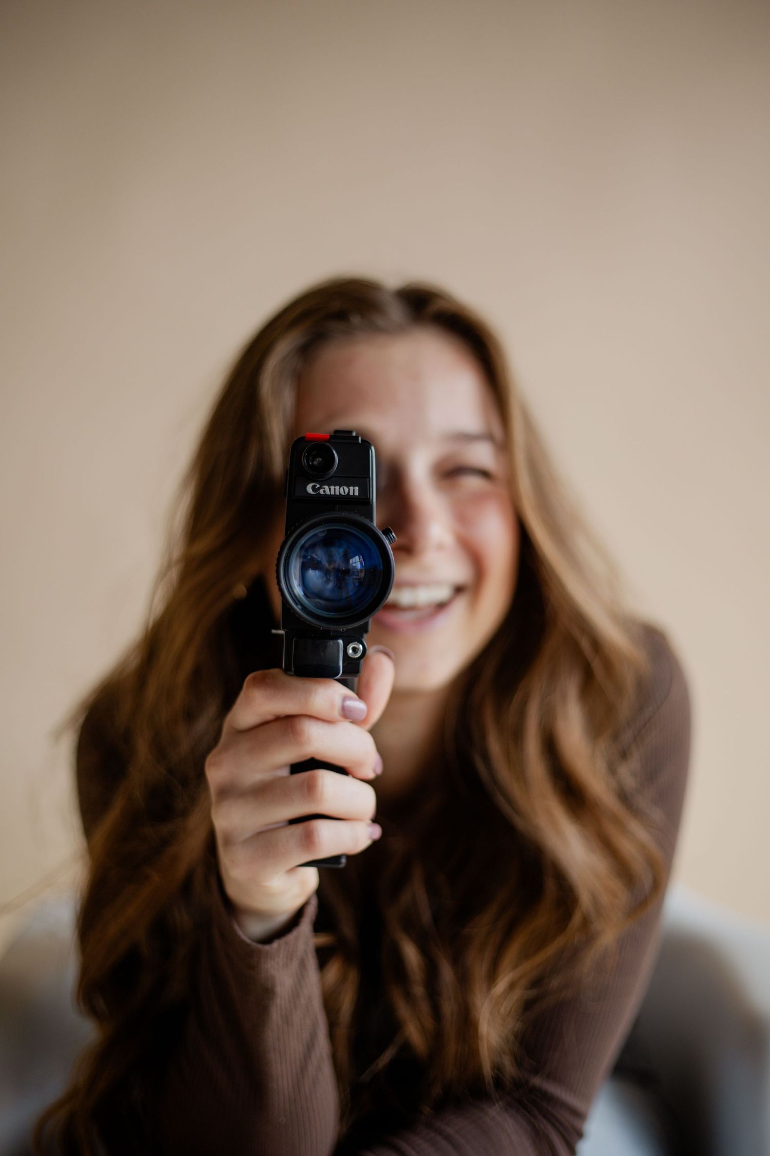Smiling young woman with long brown hair holding a Canon camera pointed forward against a neutral background.