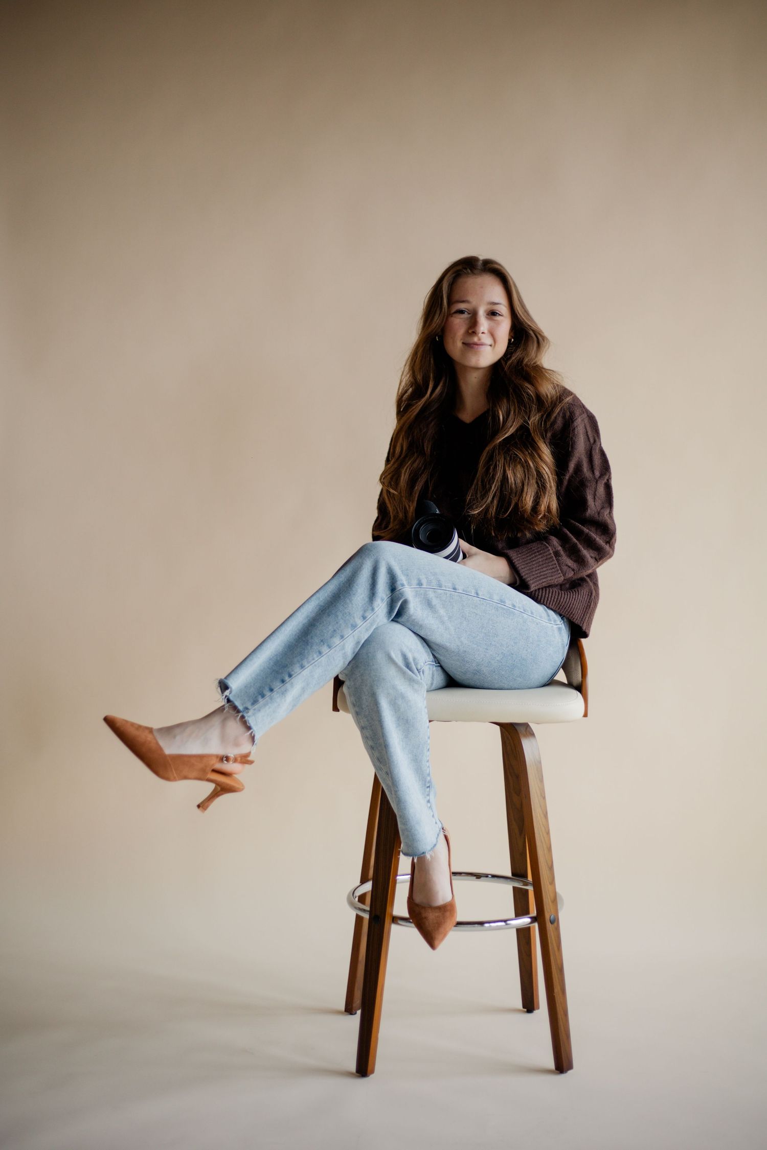 Young woman with long brown hair sitting on a wooden barstool, wearing light jeans and a dark jacket.