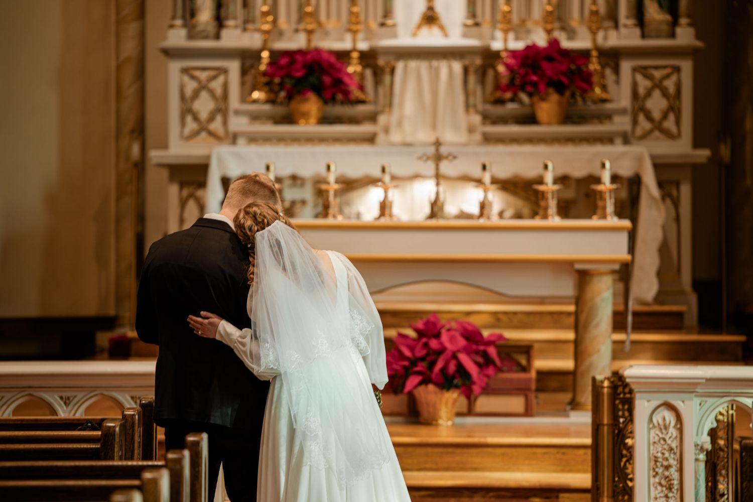 Bride and groom standing at church altar during wedding ceremony, surrounded by red poinsettias and golden decor.