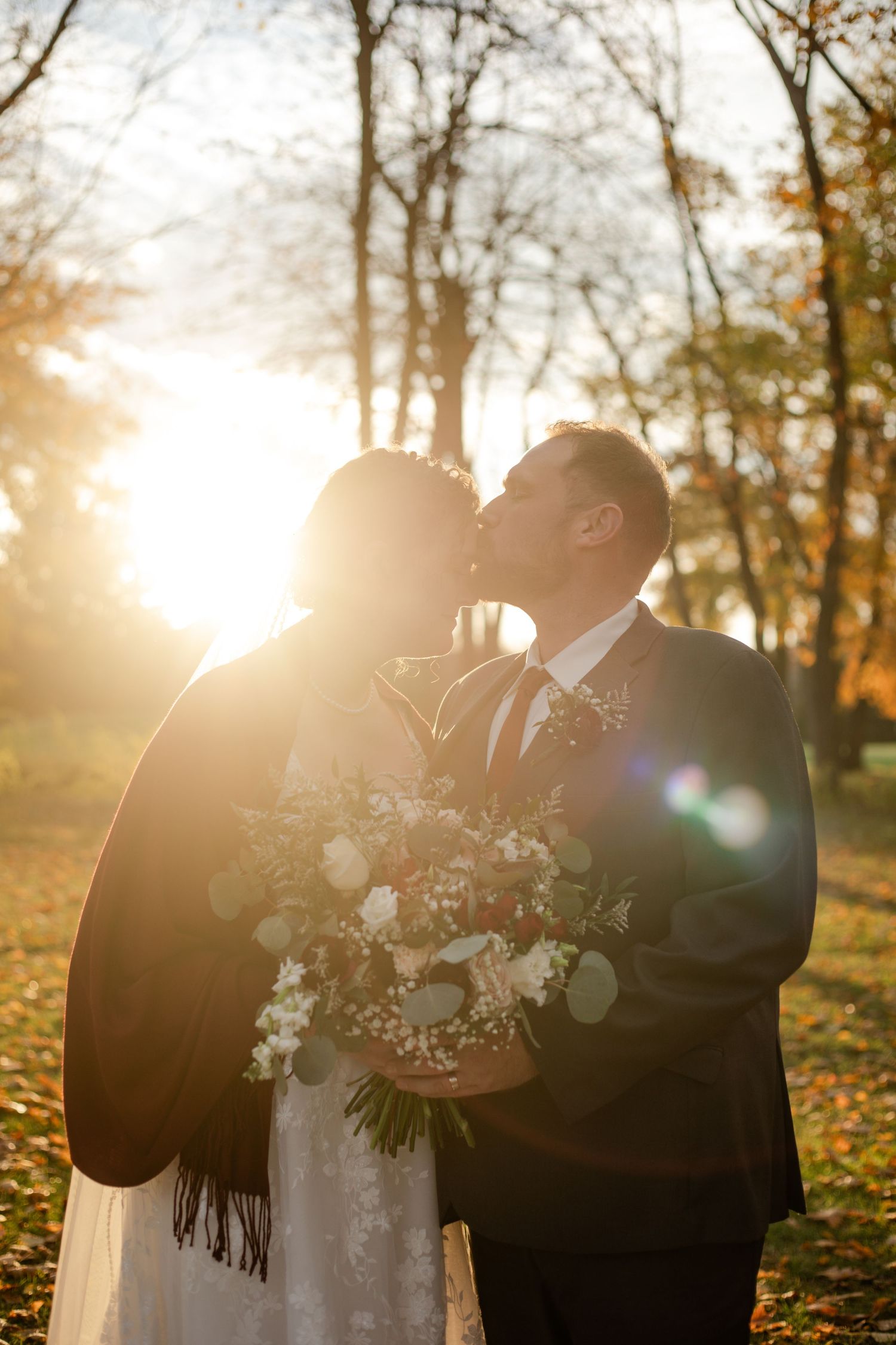 Bride and groom kissing in autumn park with golden sunset light, bride holding white floral bouquet.