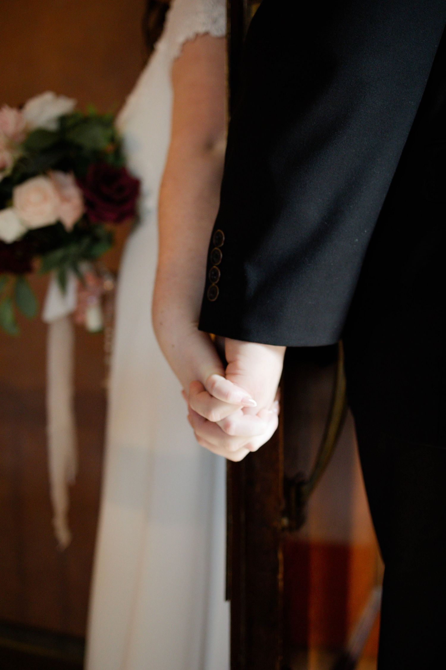 A couple in formal wedding attire holds hands in a romantic indoor setting with floral decorations visible.