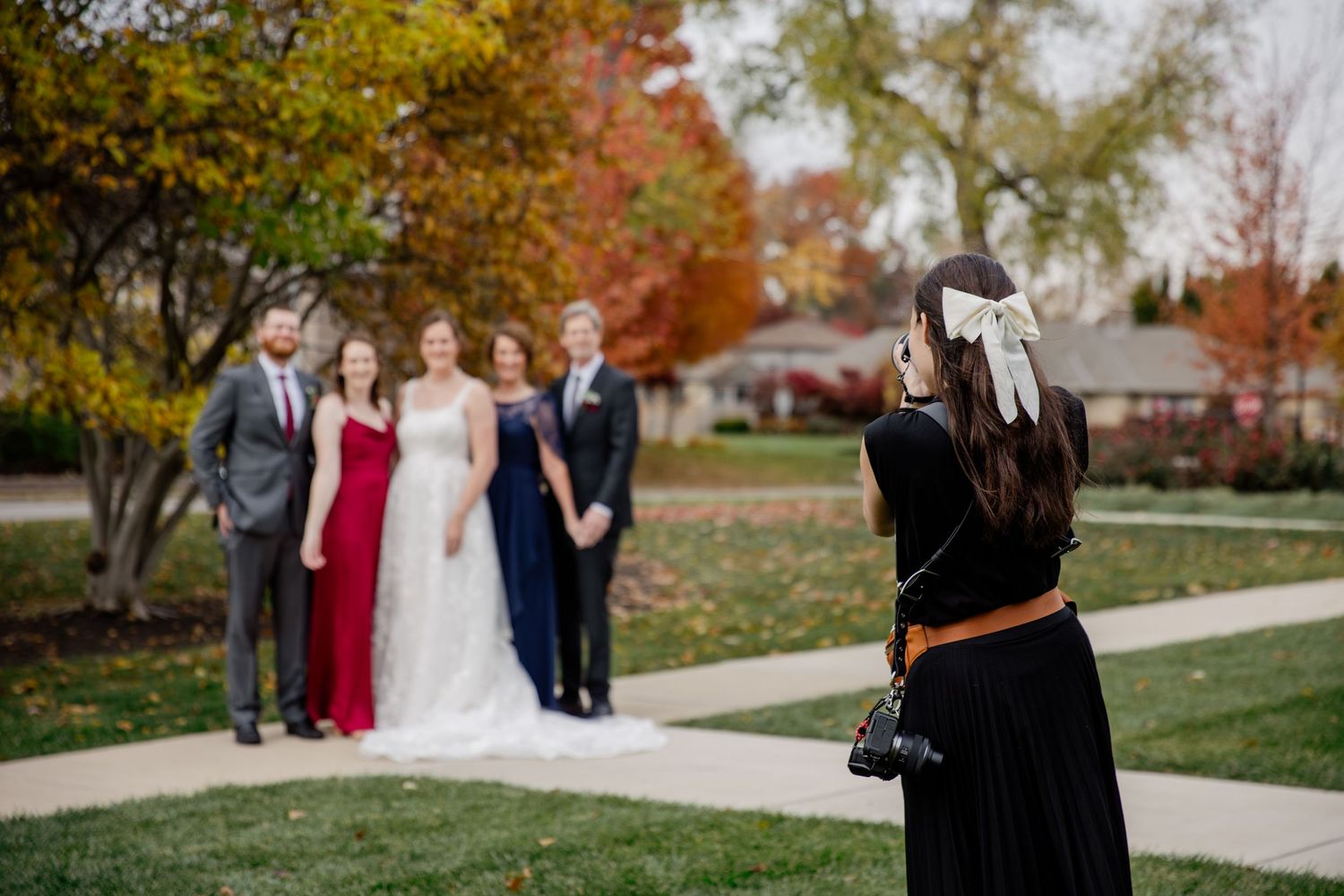 Wedding party members in formal attire gather outdoors among autumn trees and fallen leaves during a fall celebration.