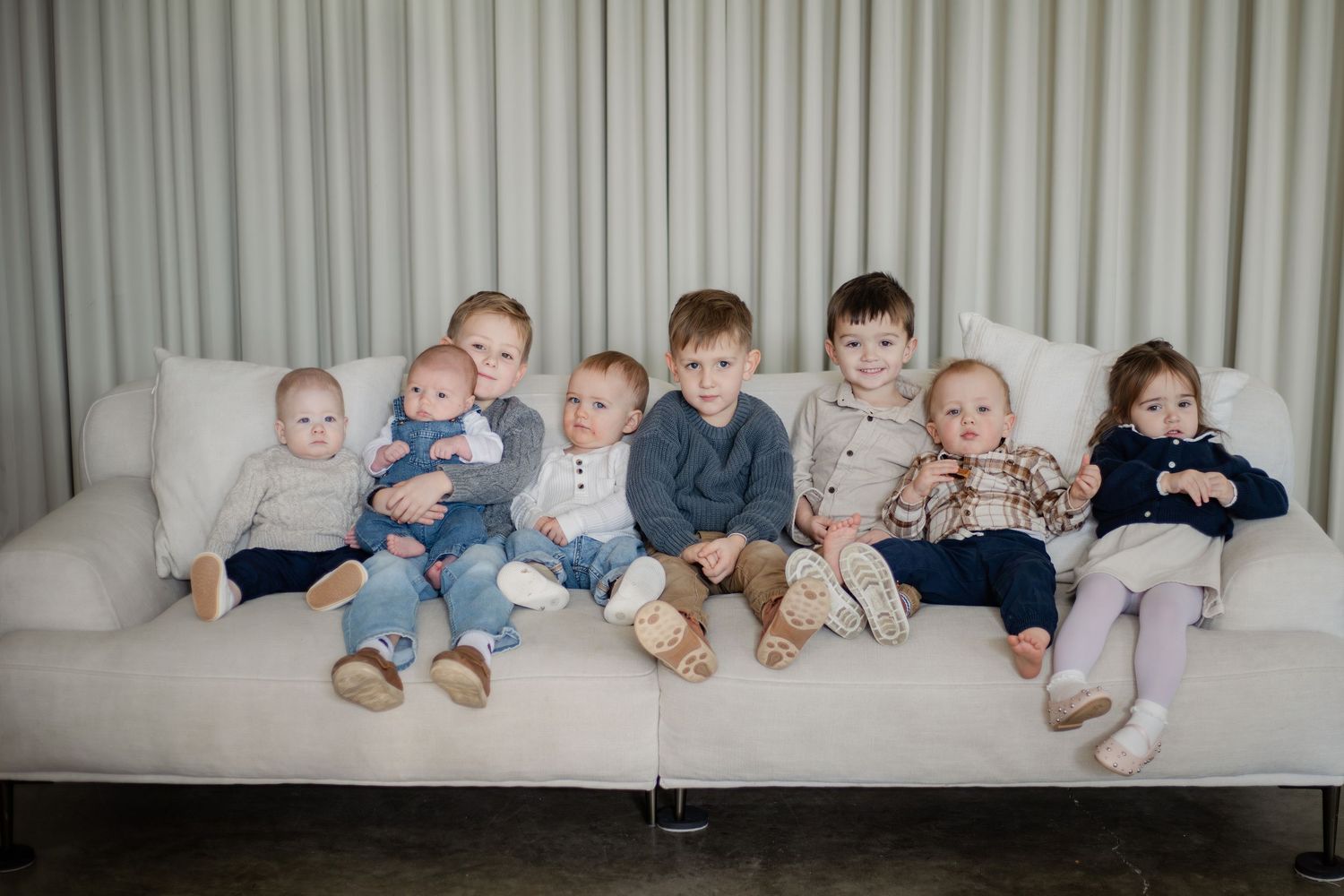 Eight young children sitting together on a white sofa in front of a light curtain backdrop.