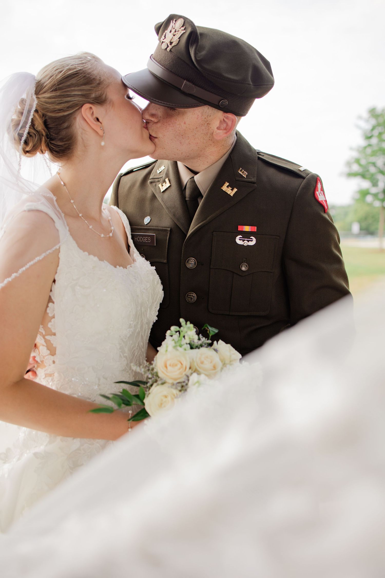 A romantic wedding moment between a bride in white and a military officer in dress uniform sharing a kiss.