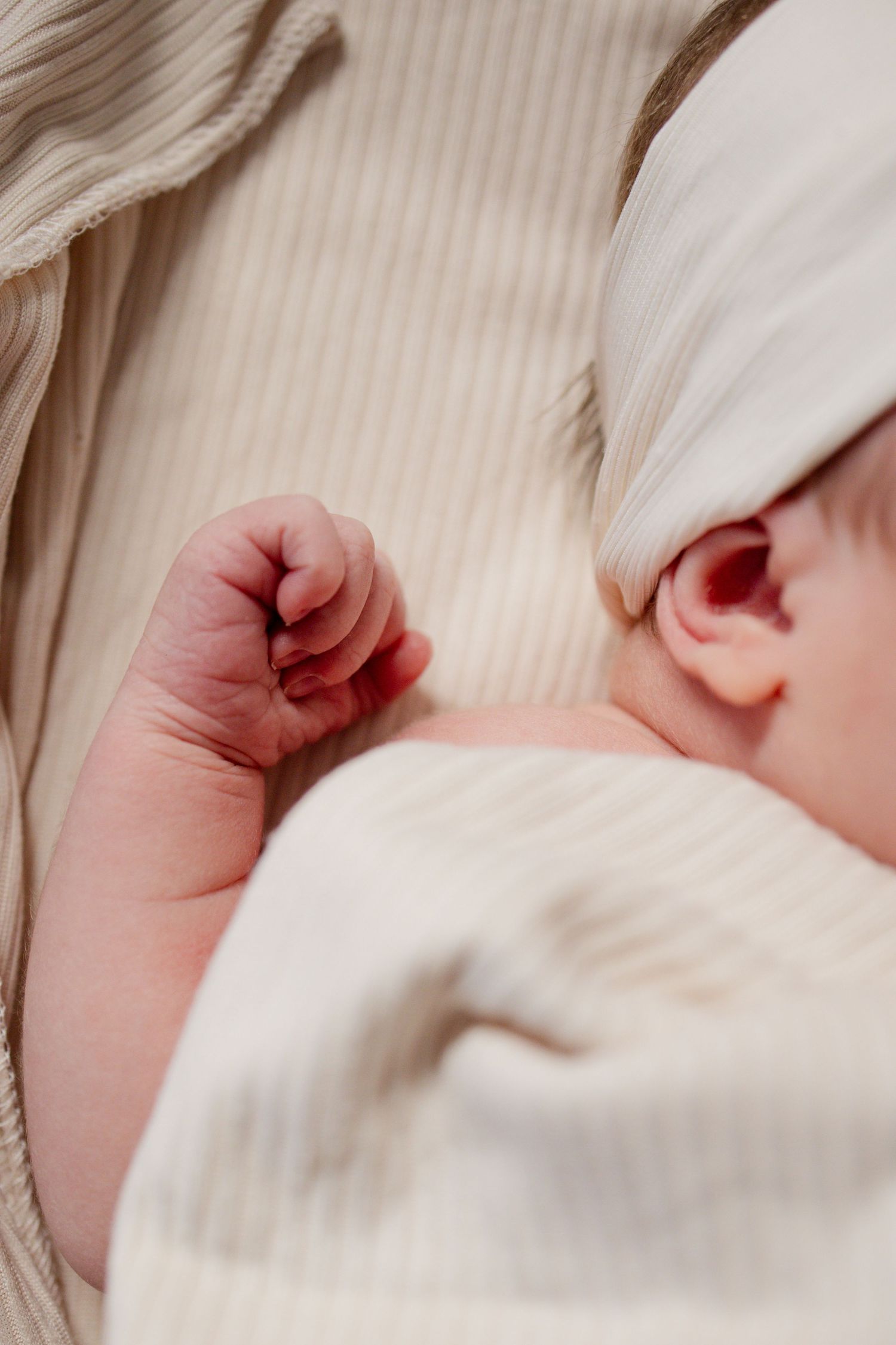 Close-up of a sleeping newborn baby's tiny fist and ear, wrapped in a soft cream knit blanket.