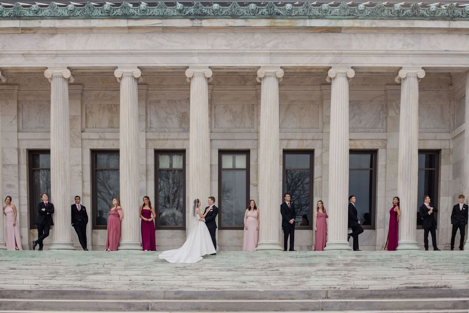 Wedding party stands elegantly posed between classic white columns of a grand building in formal attire.
