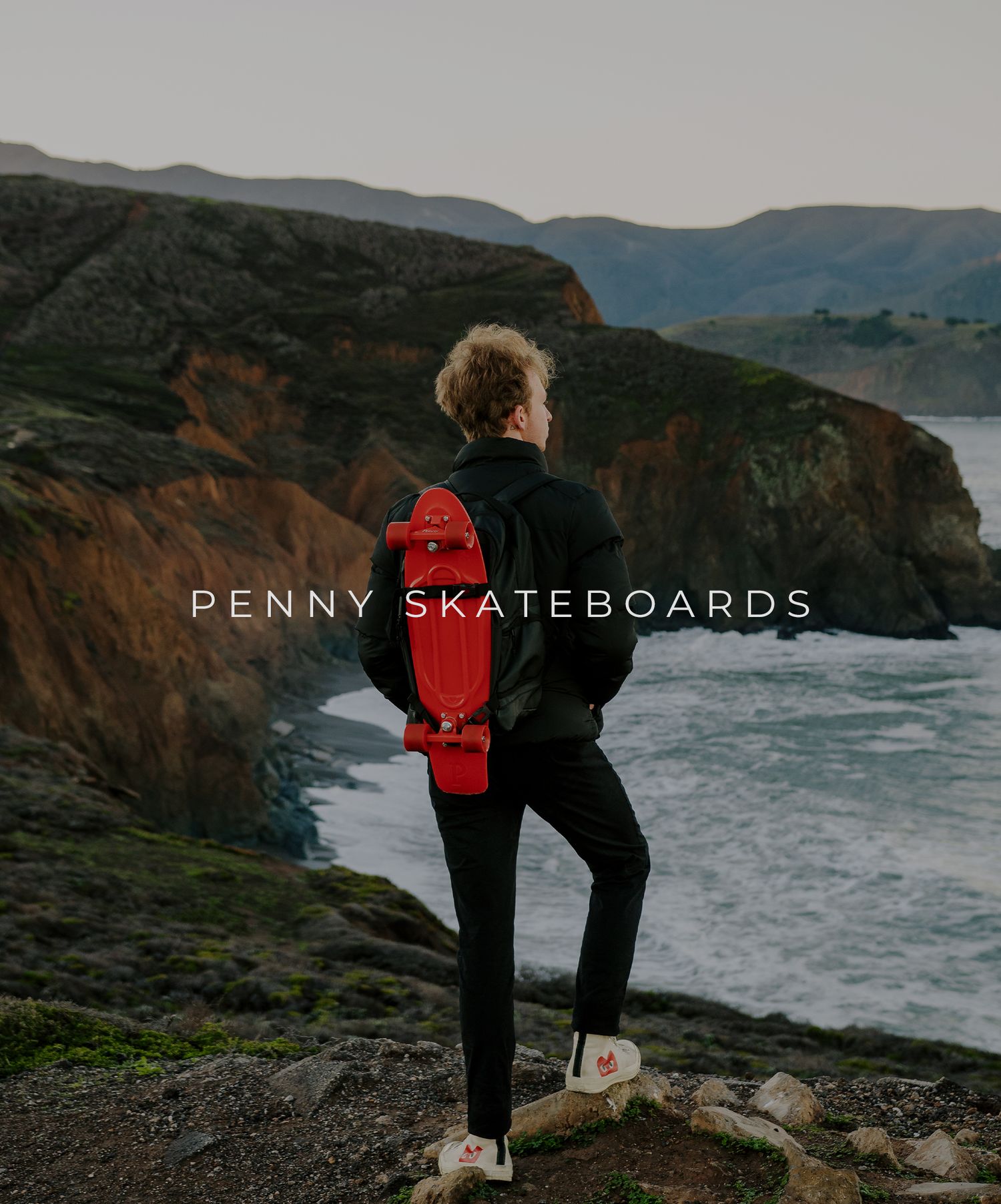 Silhouetted figure with red Penny skateboard stands on coastal cliffs overlooking dramatic rocky shoreline at sunset.