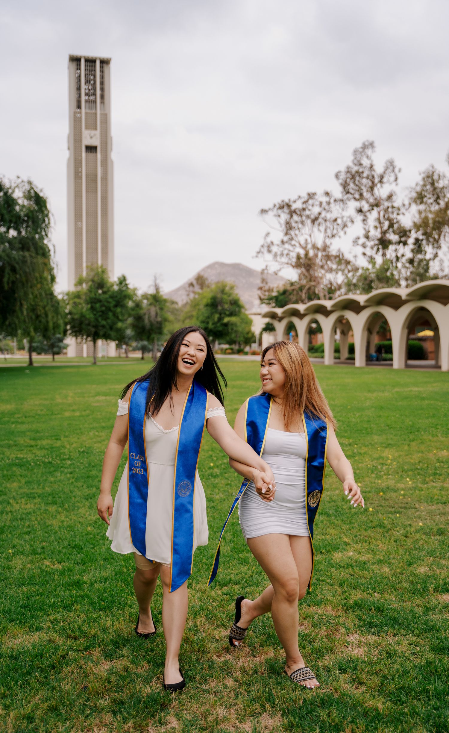 Graduates walking and laughing on campus lawn with bell tower in background.